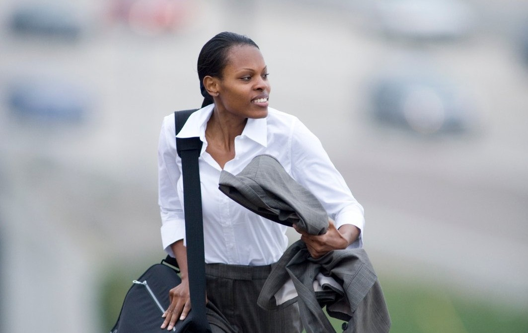 Businesswoman wearing a white shirt and gray trousers carrying a shoulder bag and jacket while walking outdoors.