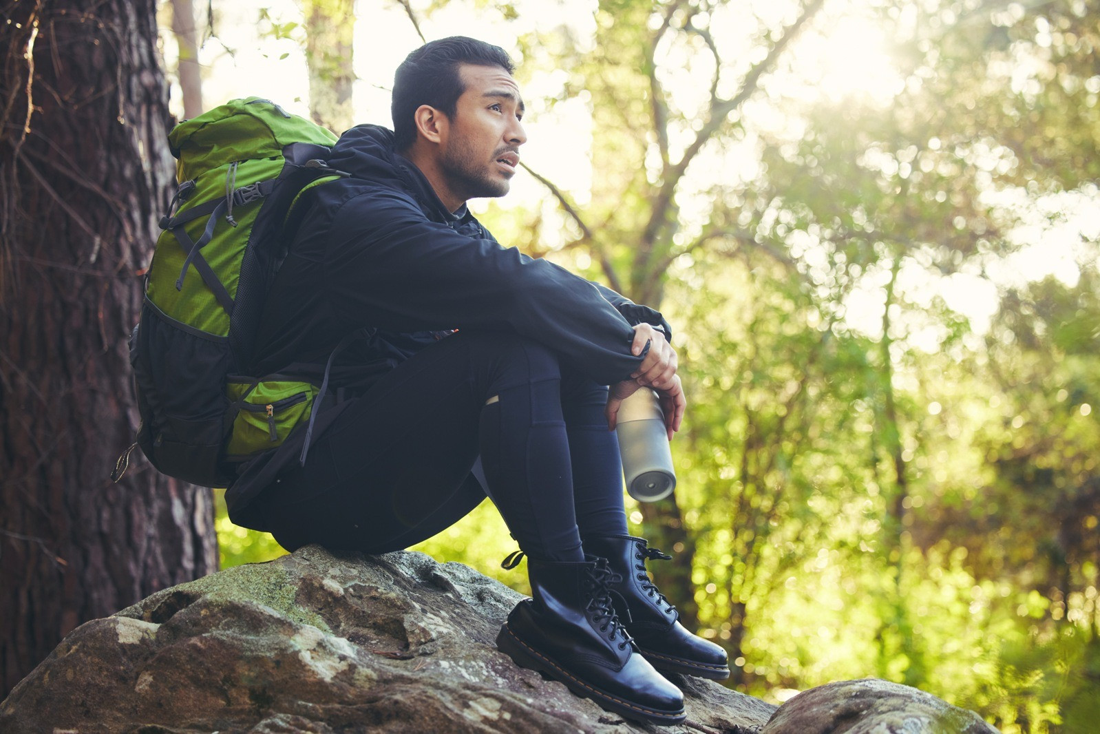 Man with a green backpack sitting on a rock in a peaceful forest, holding a water bottle and looking thoughtfully into the distance.