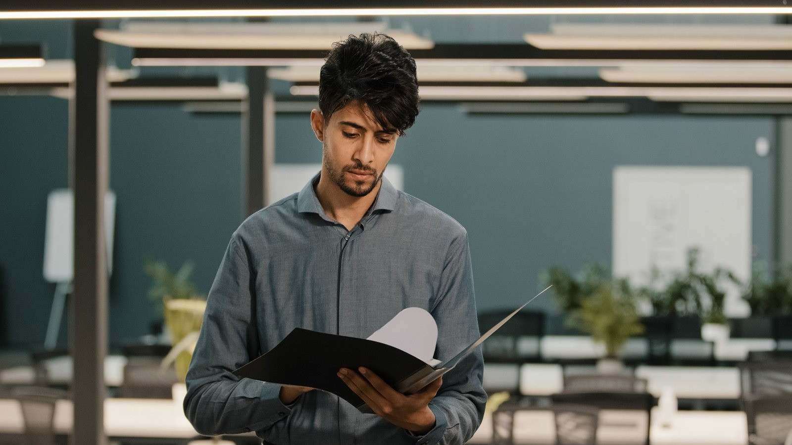 Man wearing a gray button-down shirt reviewing documents in a folder while standing in a modern office.