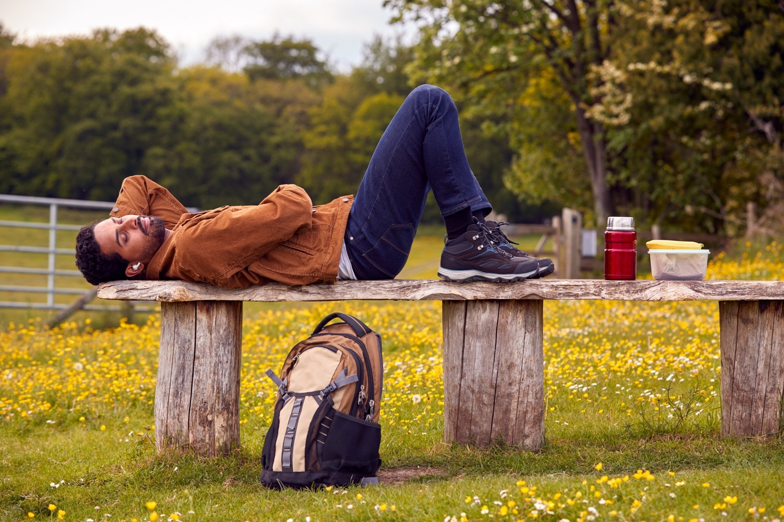 Man lying on a rustic wooden bench in a peaceful meadow filled with yellow flowers, with a backpack, water bottle, and trees nearby.