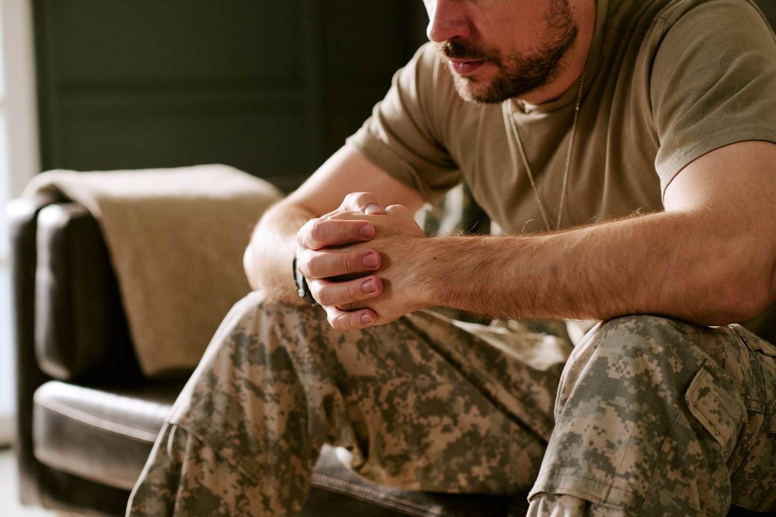Man in military clothing sitting indoors with hands clasped, representing PTSD and trauma recovery.