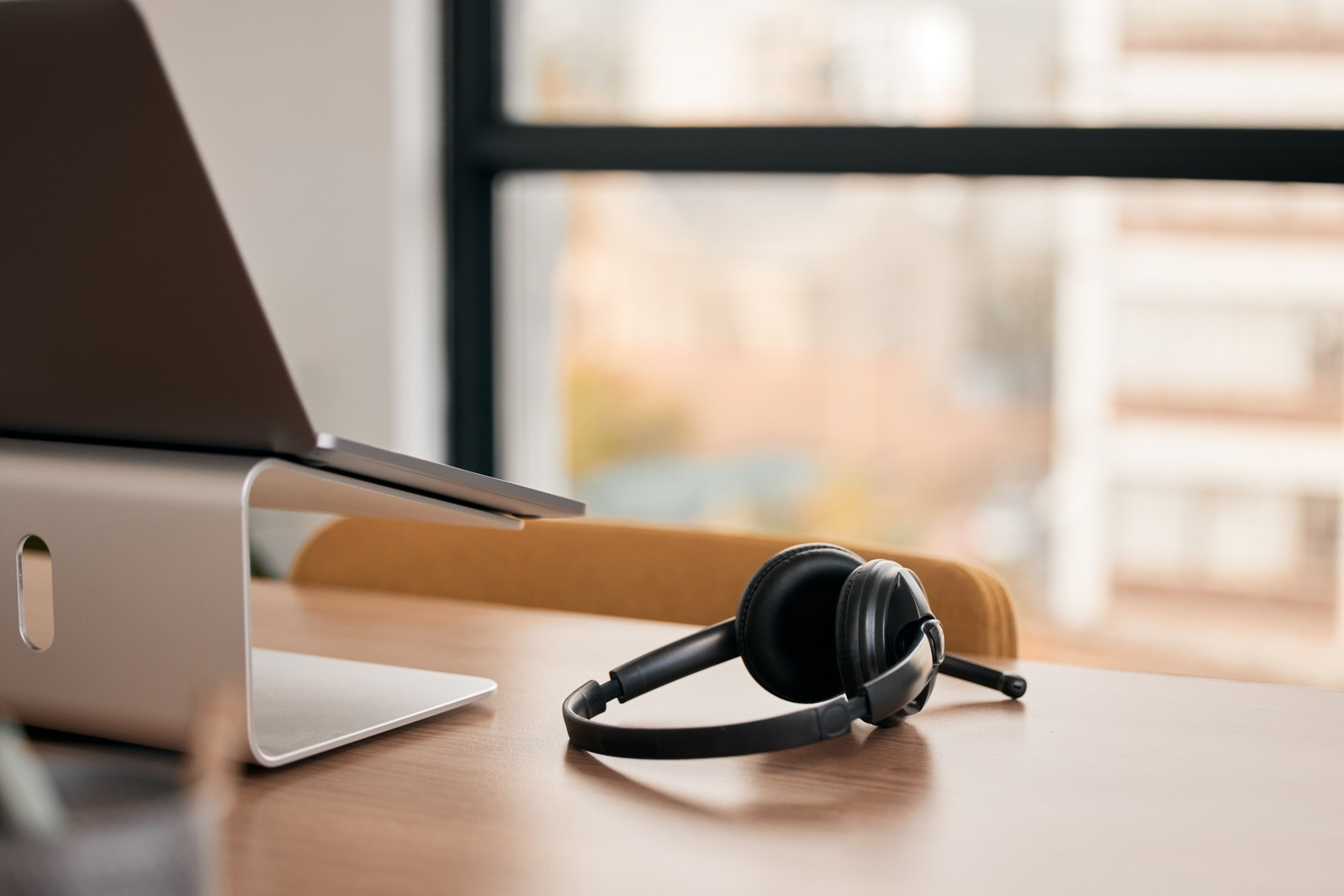 Laptop and headset resting on a desk near a window in a calm workspace.