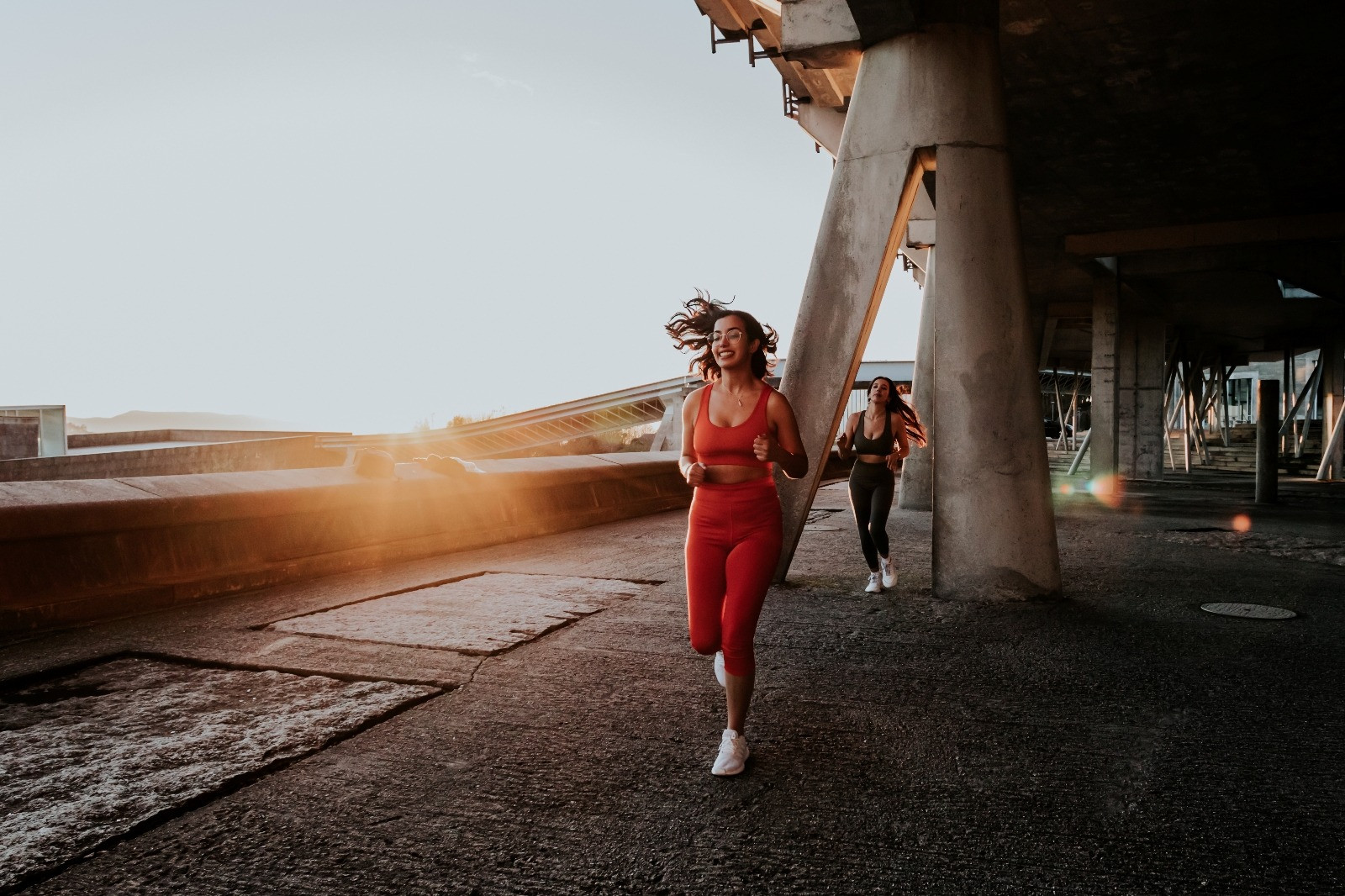 Two women jogging beside concrete supports in warm evening sunlight, with the woman in front wearing red athletic clothing.