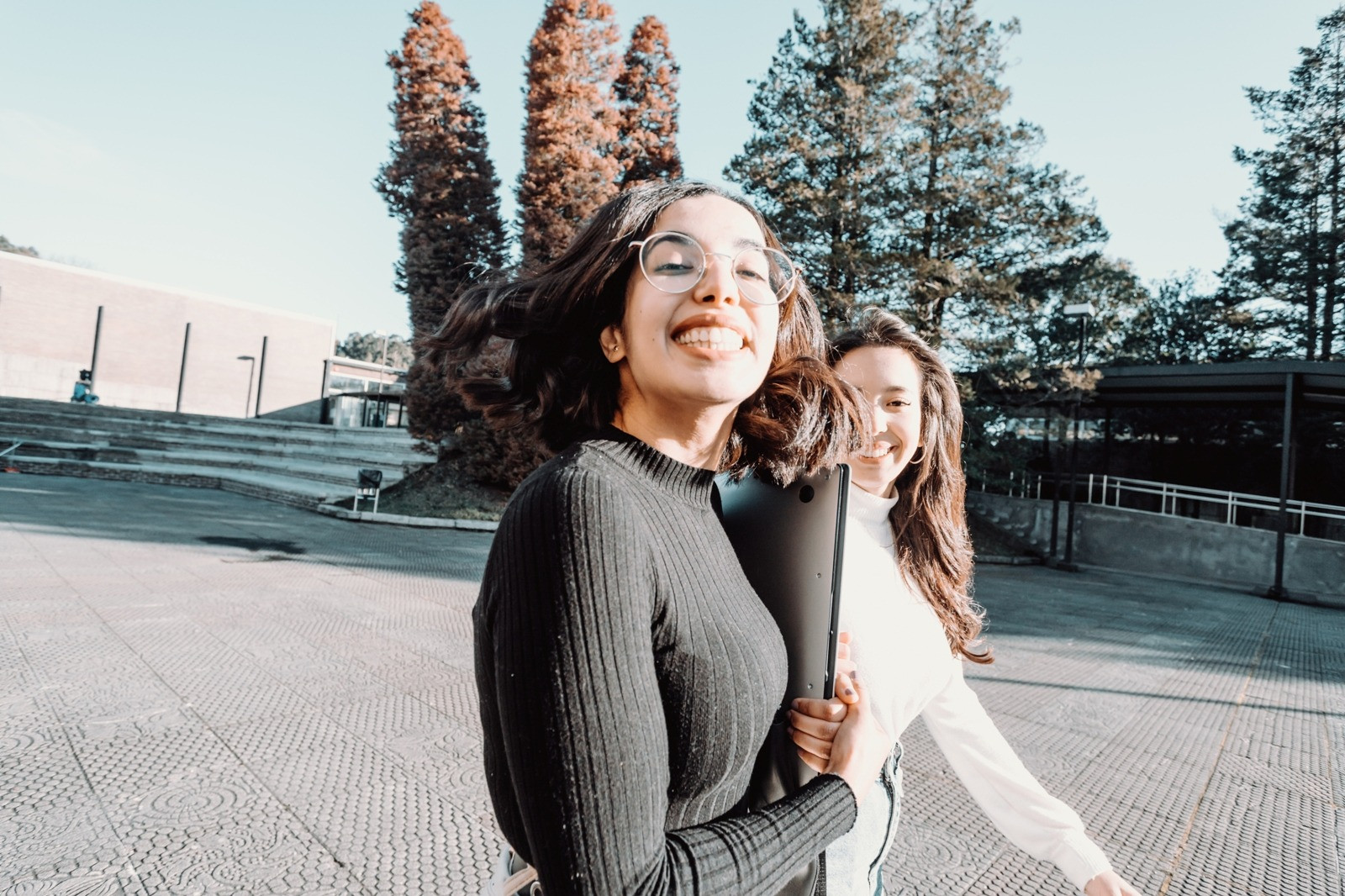 Two smiling young women walking outdoors on a sunny day, with one wearing glasses and holding a laptop against a background of trees and buildings.
