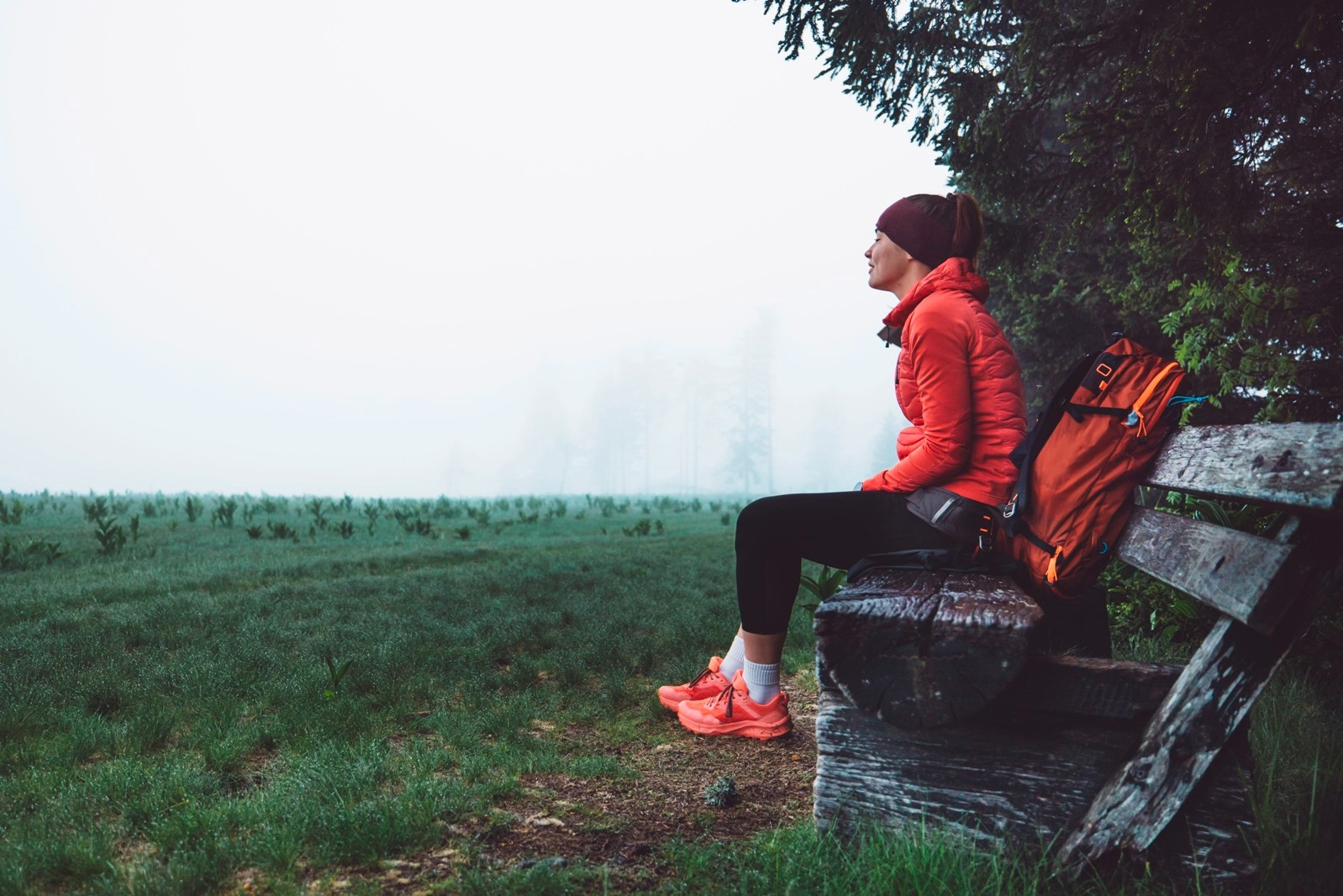 A woman in a red jacket sitting on a wooden bench beside a backpack, overlooking a misty meadow.