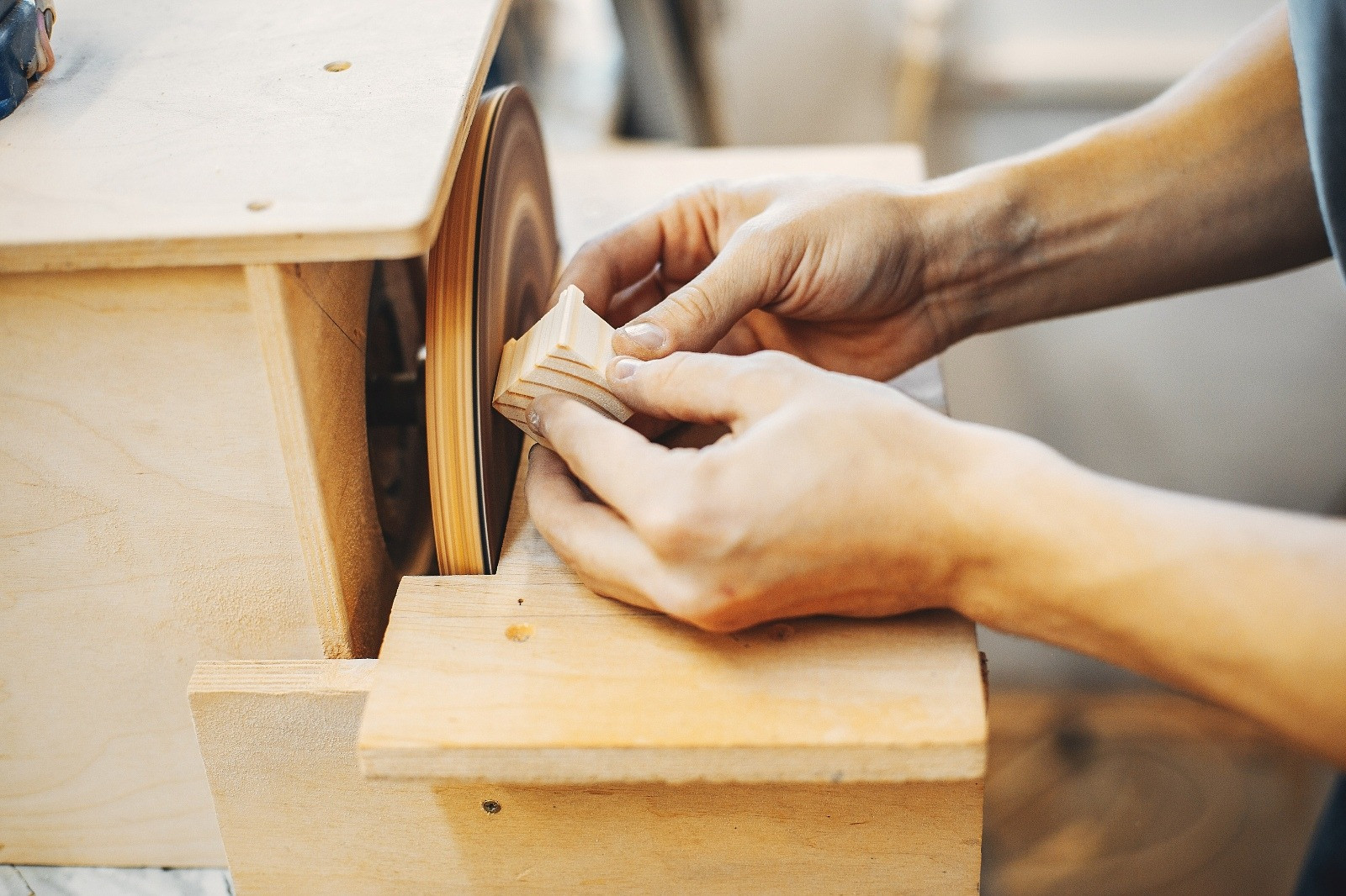 Close-up of a person carefully shaping a small wooden piece against a sanding wheel in a woodworking workshop.