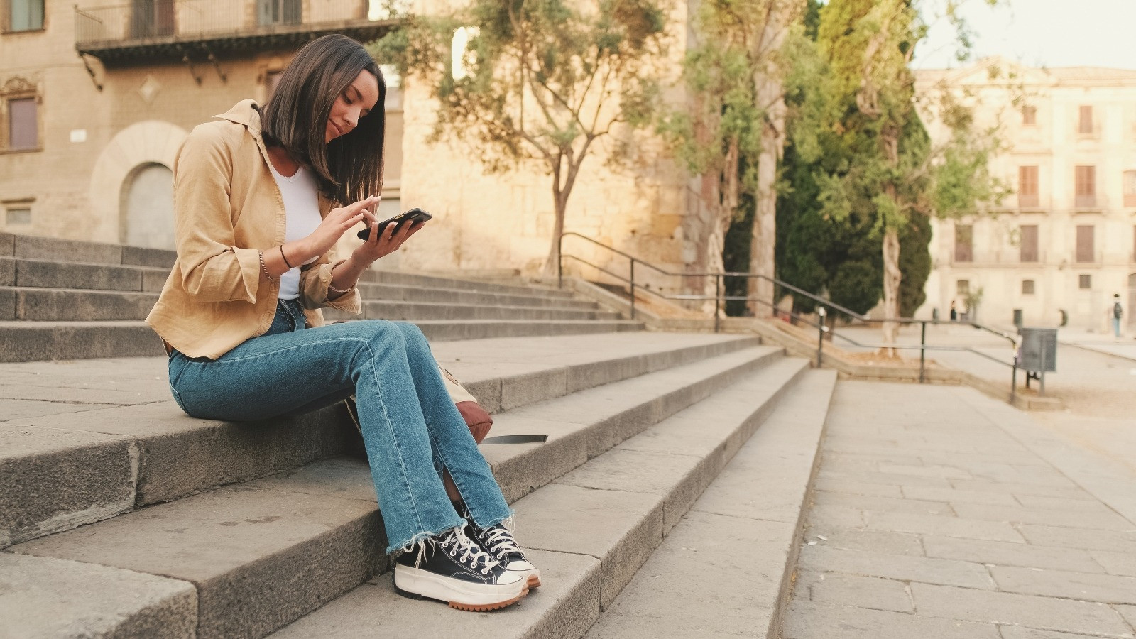 Woman sitting on outdoor stone steps while using her smartphone, representing the convenience of exploring online EMDR tools and programs.
