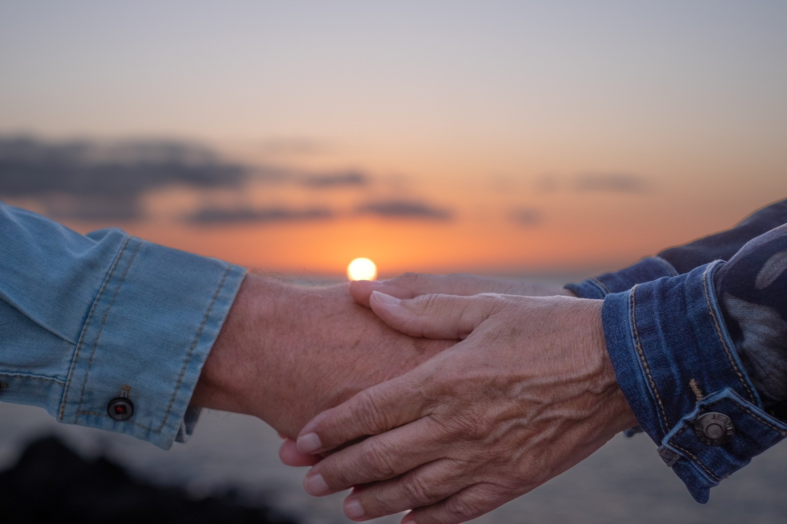 Two people holding hands against a sunset sky, suggesting quiet support and connection.
