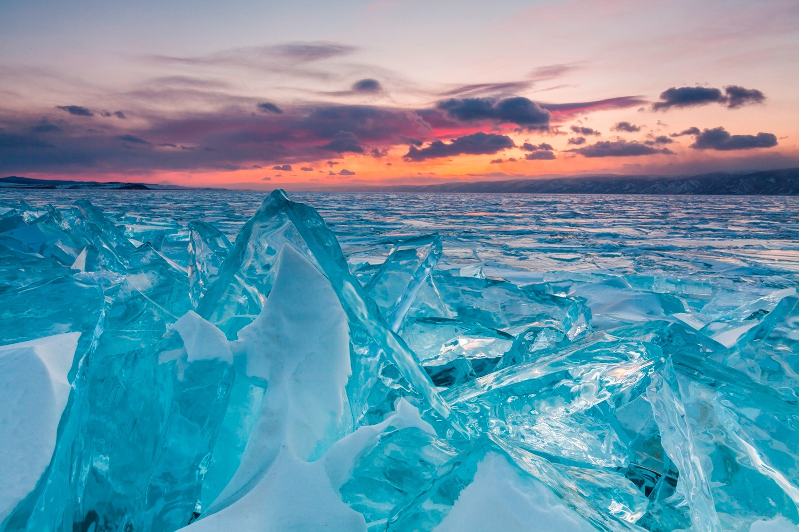 Jagged blue ice formations on a frozen lake beneath a pink and orange sunset sky.