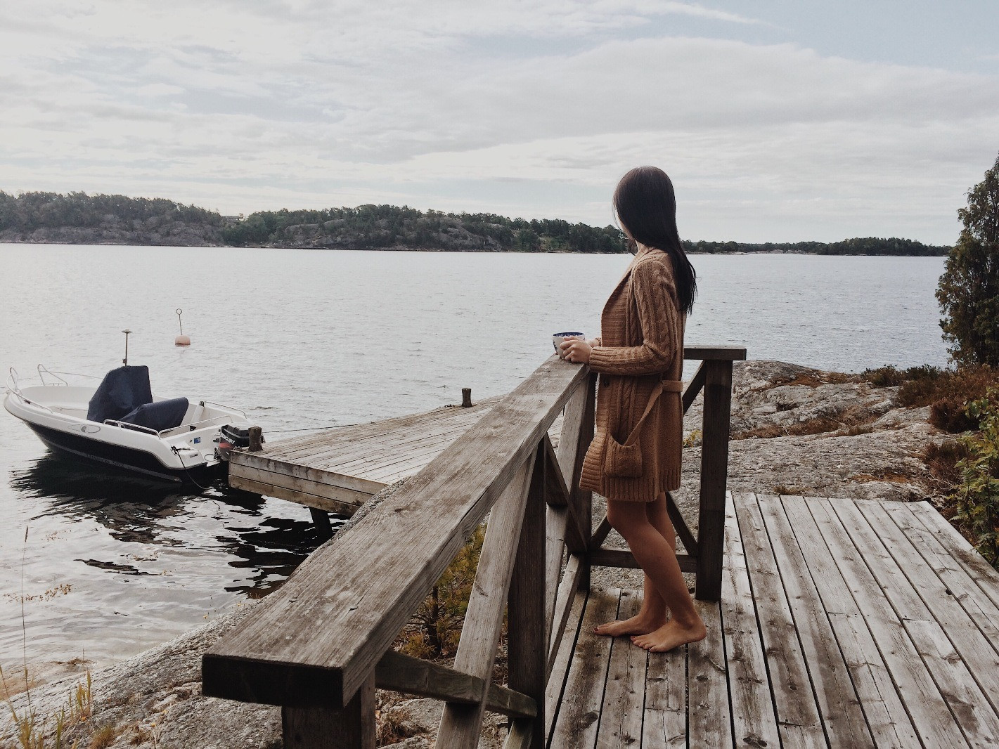 A woman in a brown cardigan standing barefoot on a wooden deck, holding a cup and looking out over a peaceful lake.