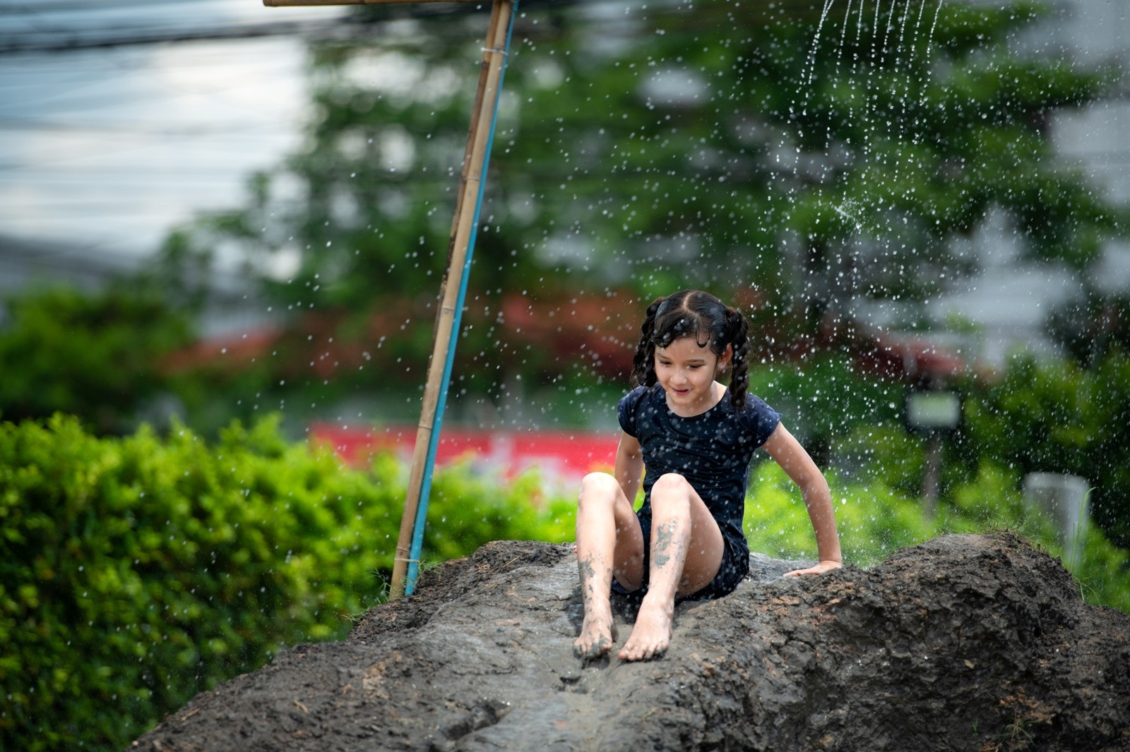 Young girl sitting on a muddy mound under falling water, smiling while enjoying outdoor play in a green garden.