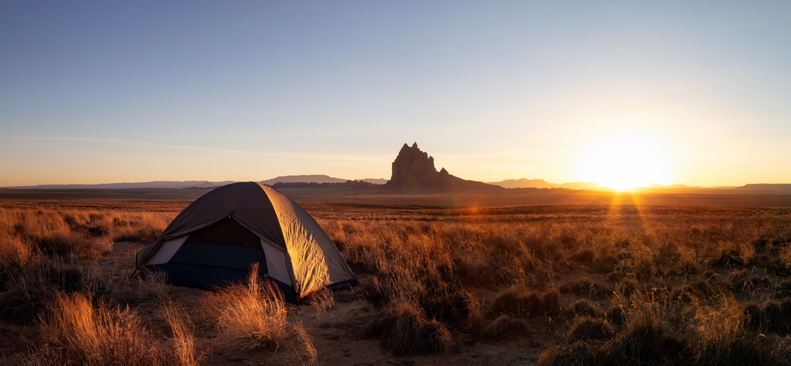 Tent set up in an open landscape at sunset, with warm light across the grass and distant rock formations.
