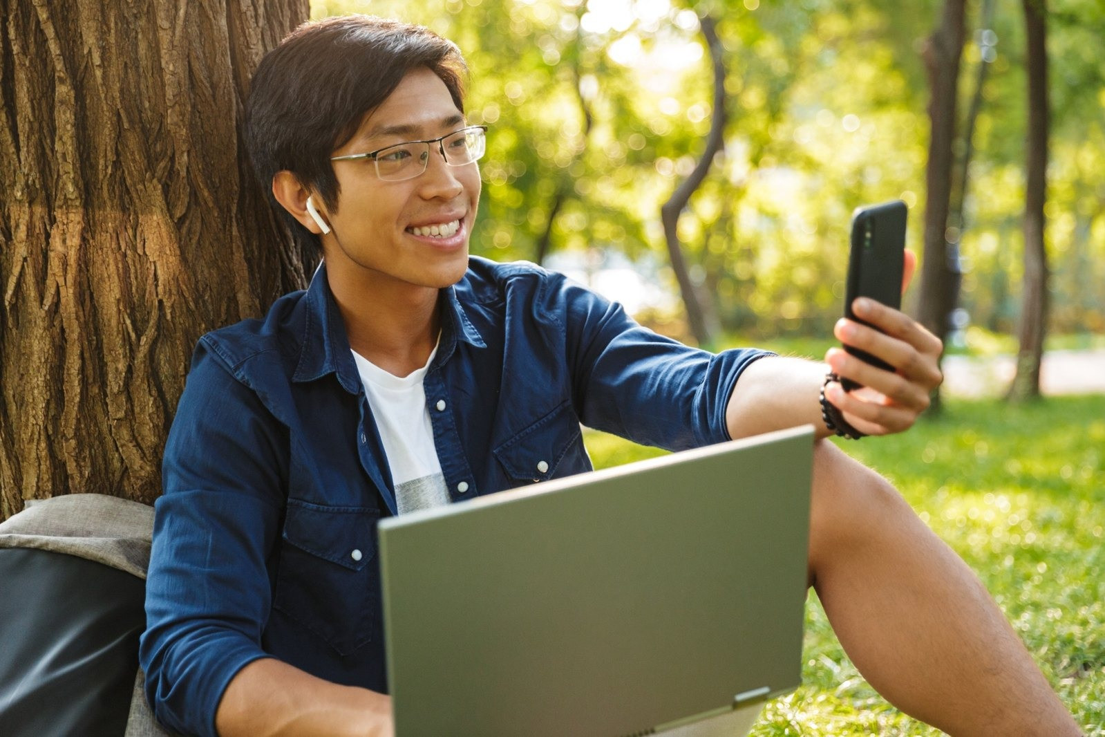 Smiling man wearing glasses and earbuds while sitting against a tree outdoors, using a smartphone with a laptop on his lap.