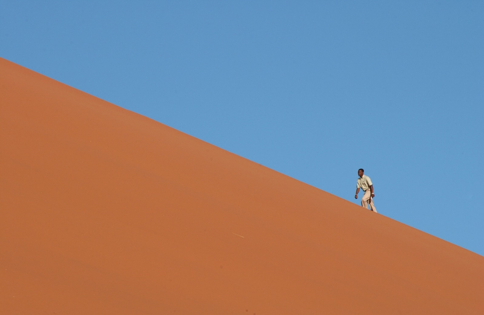A man walking up an orange sand dune beneath a clear blue sky.
