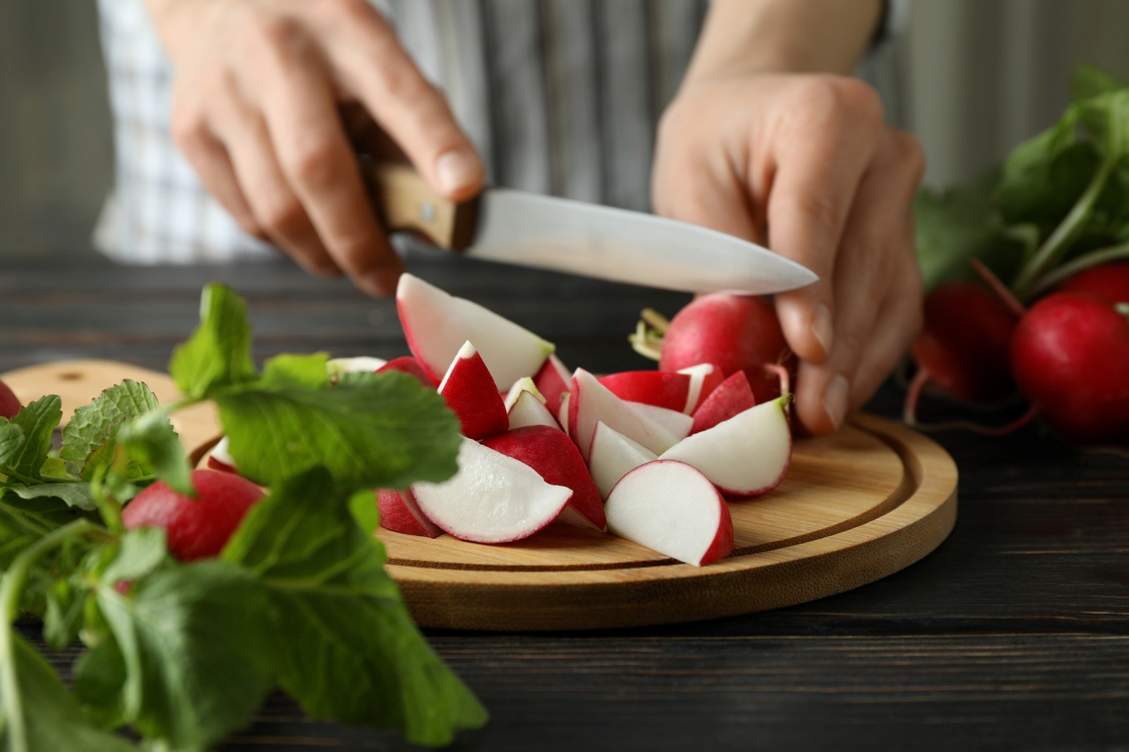 Hands slicing fresh radishes on a wooden cutting board.