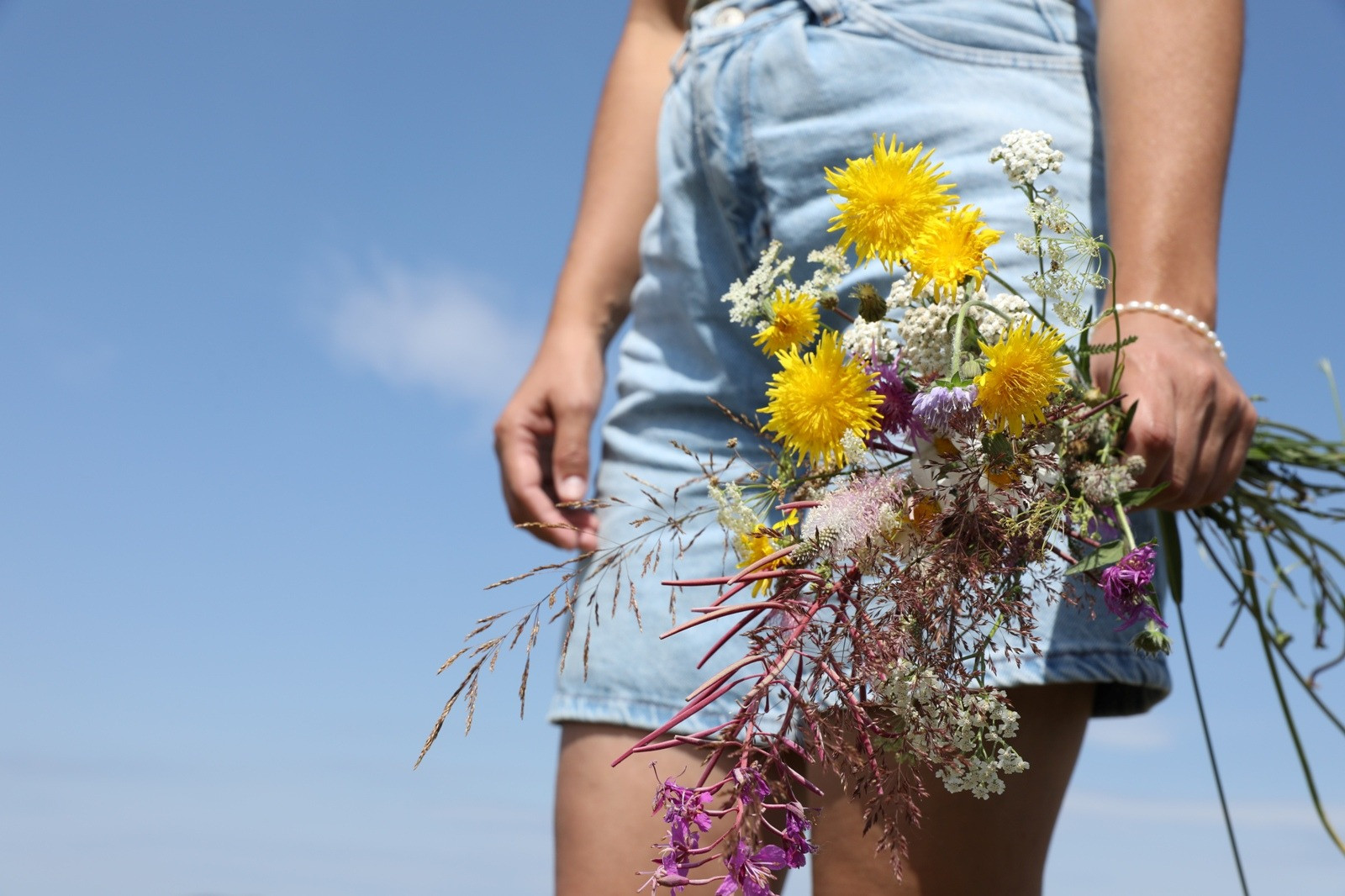 Person in a light blue denim outfit holding a colorful bouquet of yellow, purple, and white wildflowers against a clear blue sky.