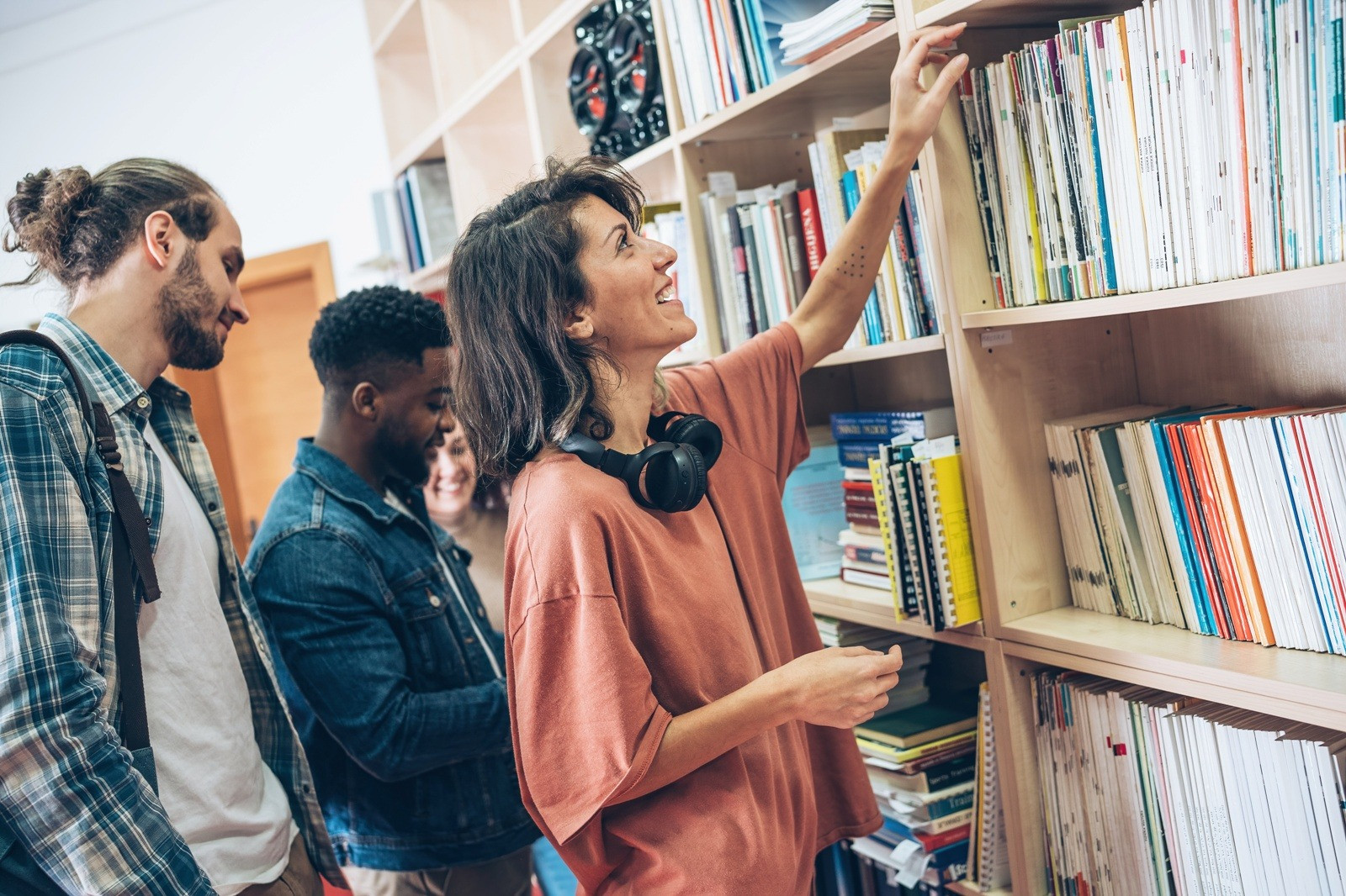 Group of people browsing books in a library, with a woman wearing headphones around her neck reaching for a book on a shelf.