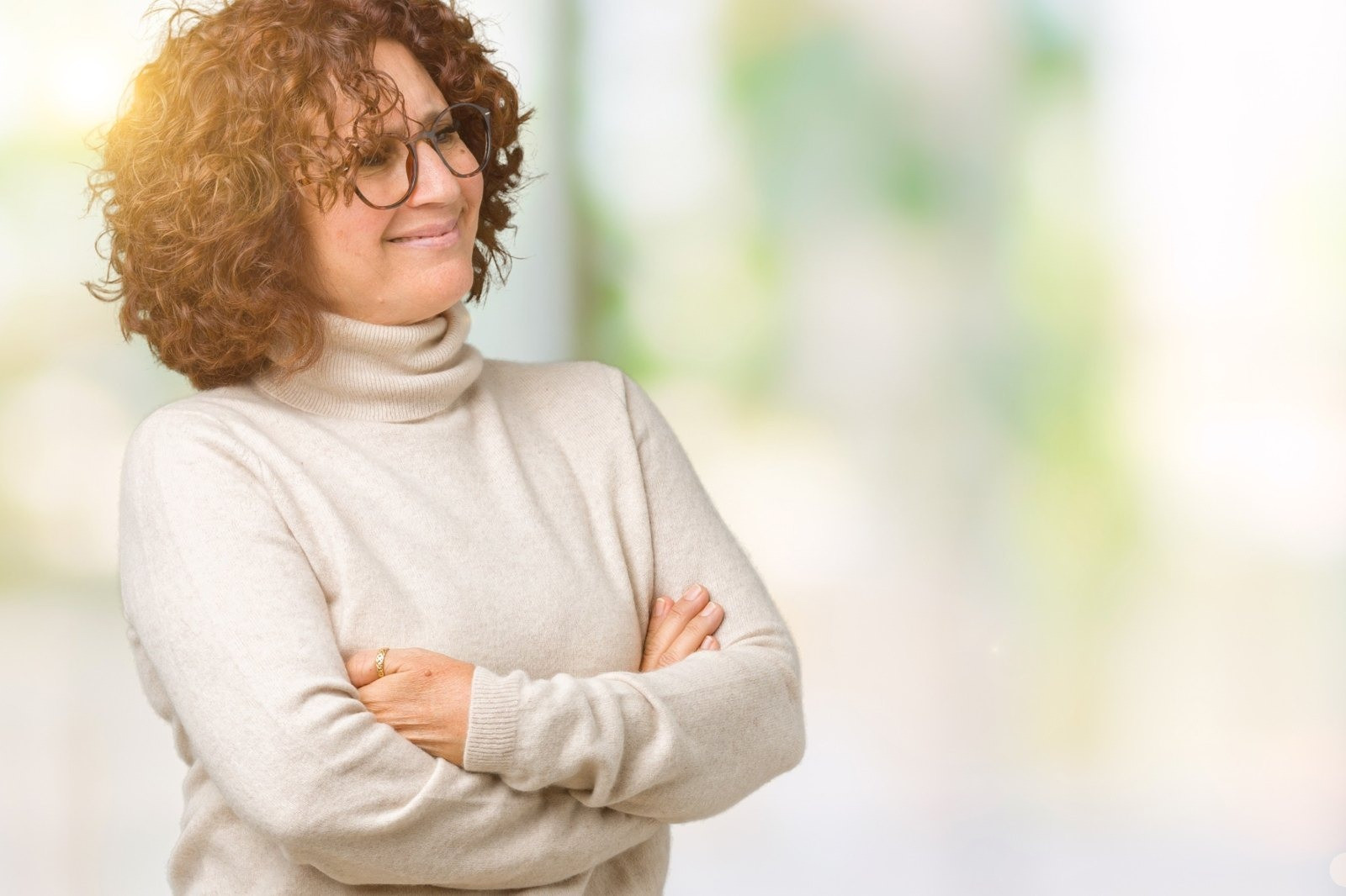Smiling woman with curly red hair and glasses wearing a cream turtleneck sweater with her arms crossed.