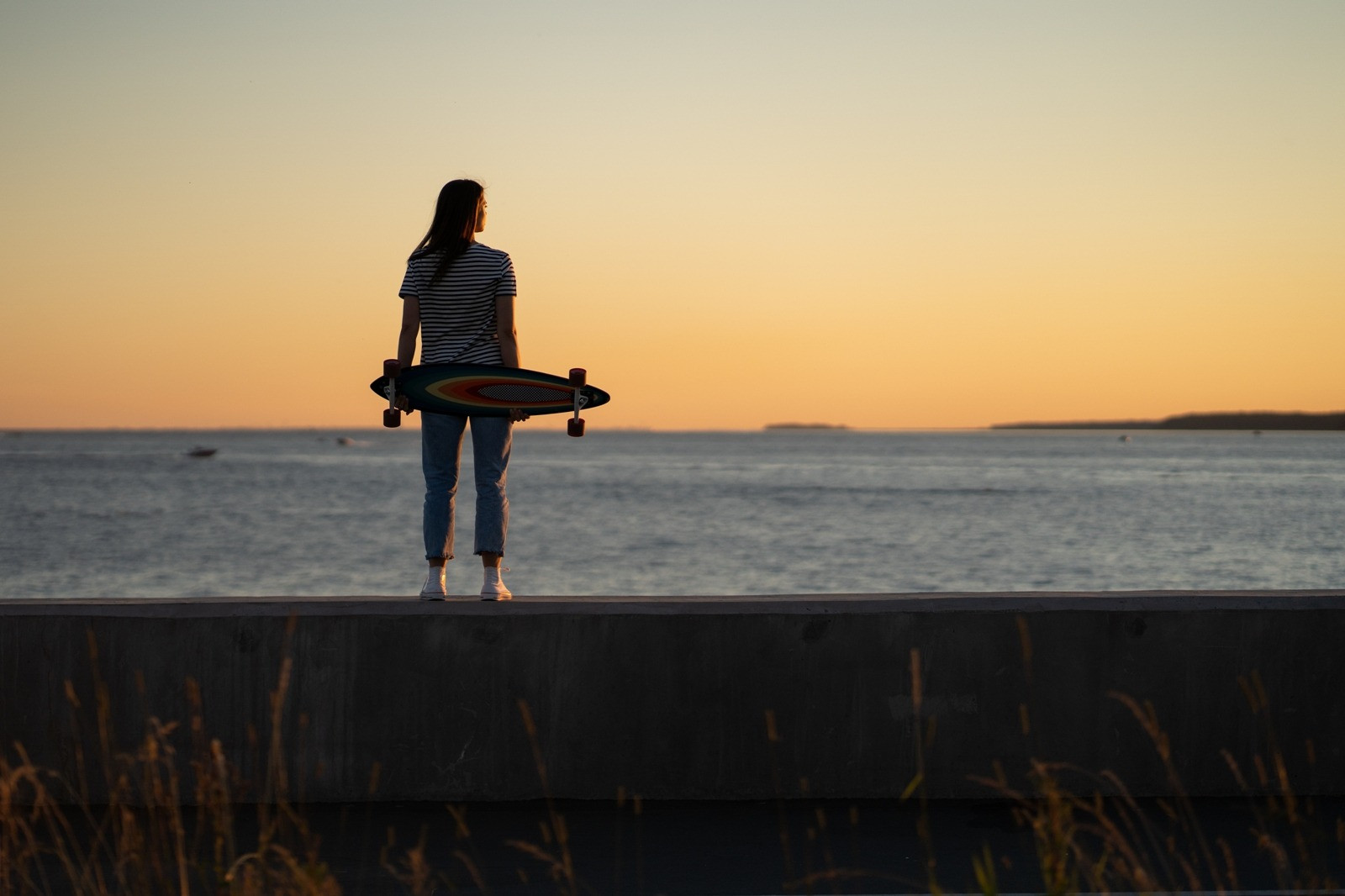 Woman holding a skateboard while standing by the ocean at sunset, looking toward the horizon under a warm golden sky.
