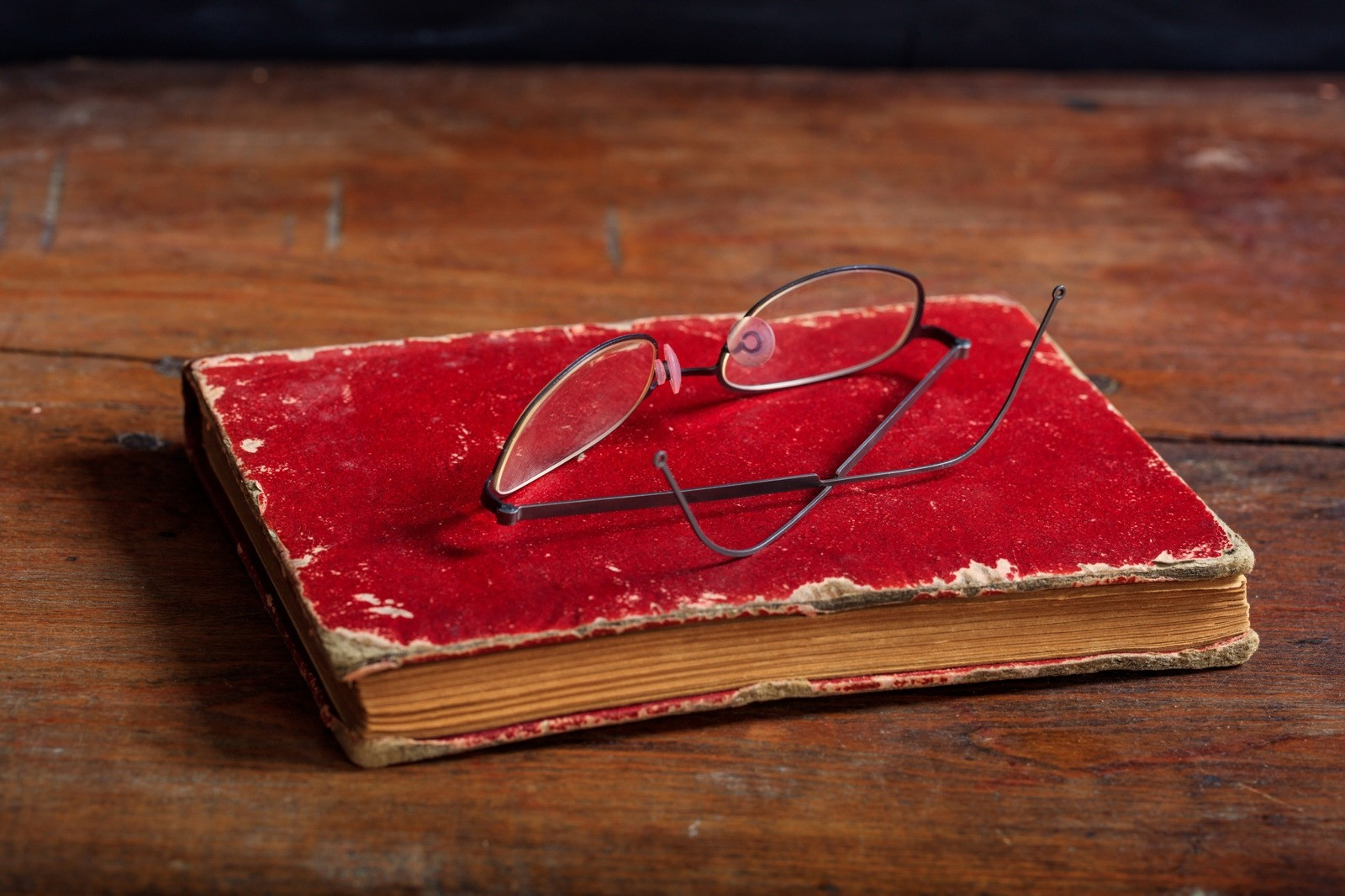 Eyeglasses resting on a worn red hardcover book on a wooden table.
