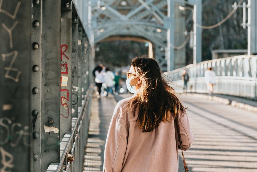 Woman wearing a face mask walking across a pedestrian bridge in daylight, with other people in the background.
