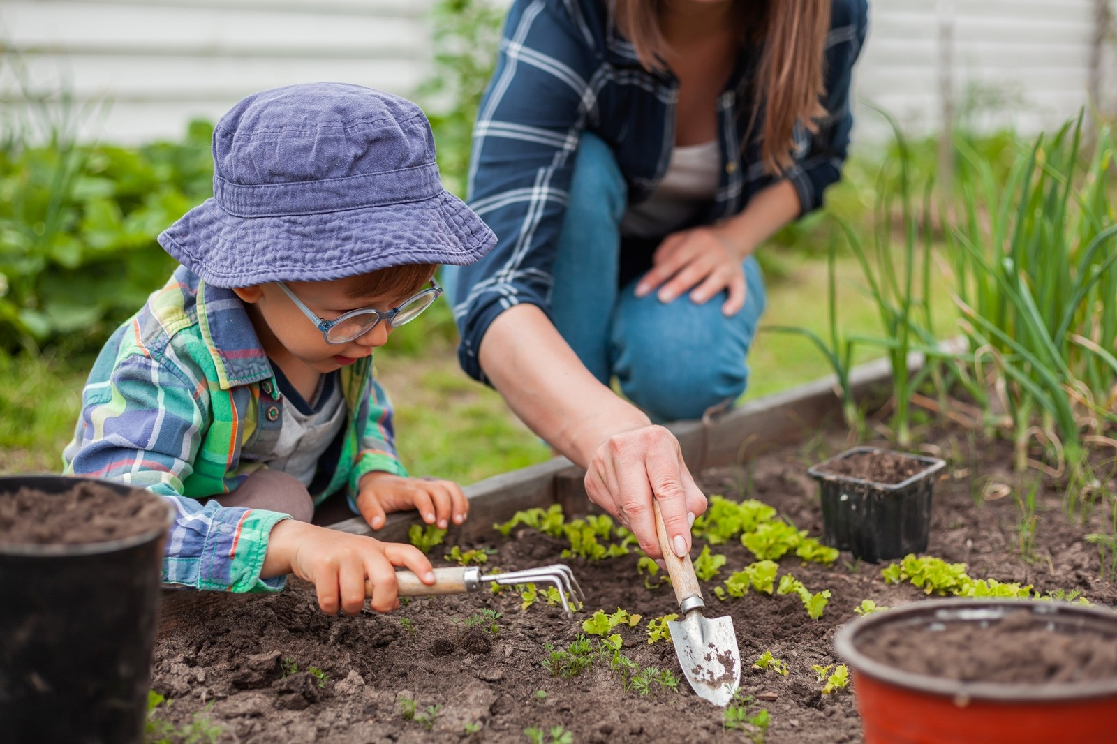 A young child wearing a blue hat and glasses gardening alongside an adult woman, using small tools to tend to plants in a garden.