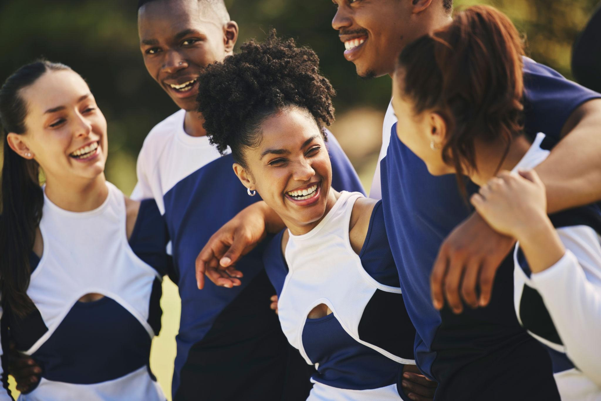 Five smiling young adults standing close together outdoors, wearing coordinated navy and white athletic clothing.