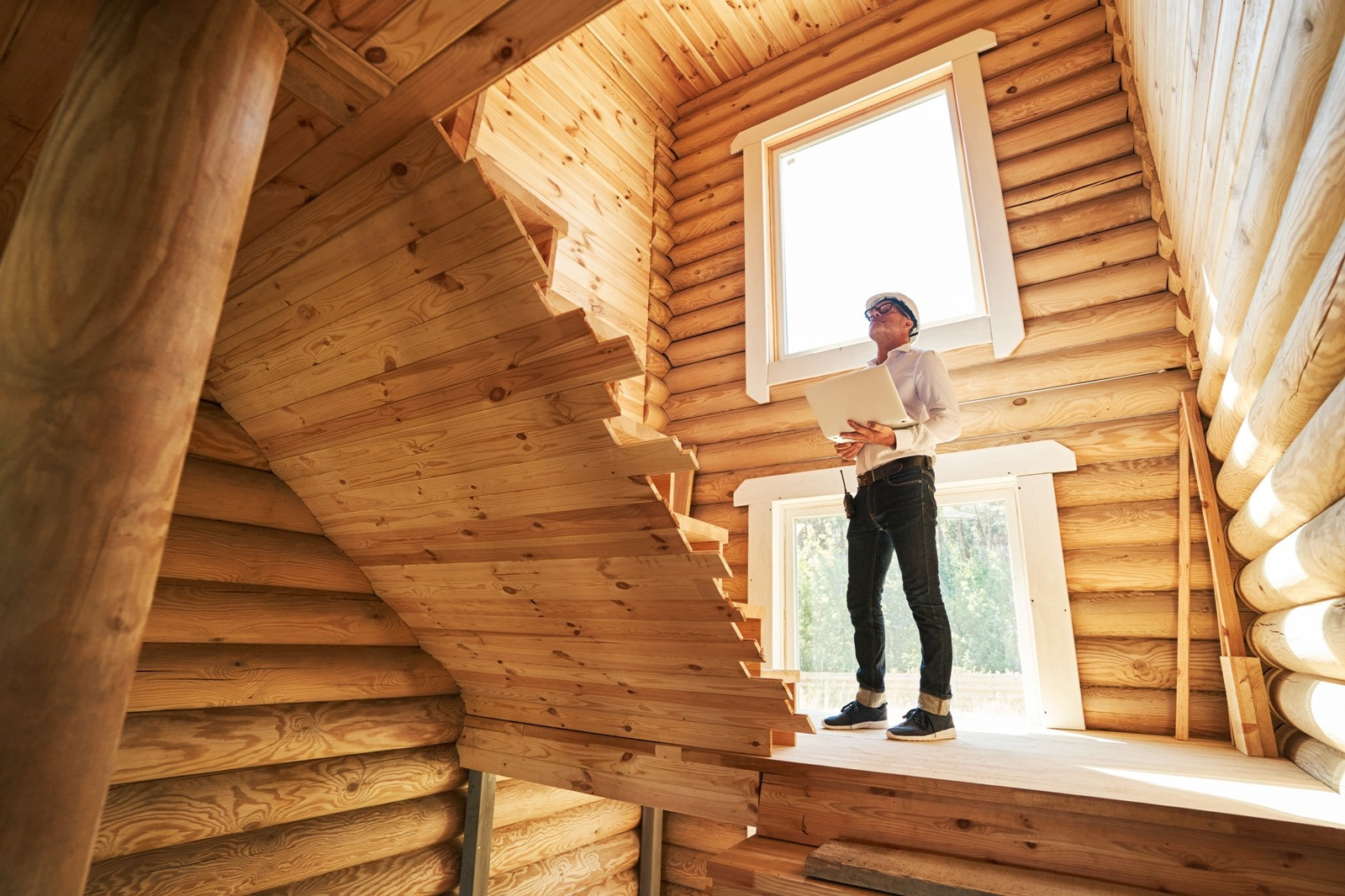 Man wearing glasses and holding papers while standing near a window inside a sunlit wooden house.