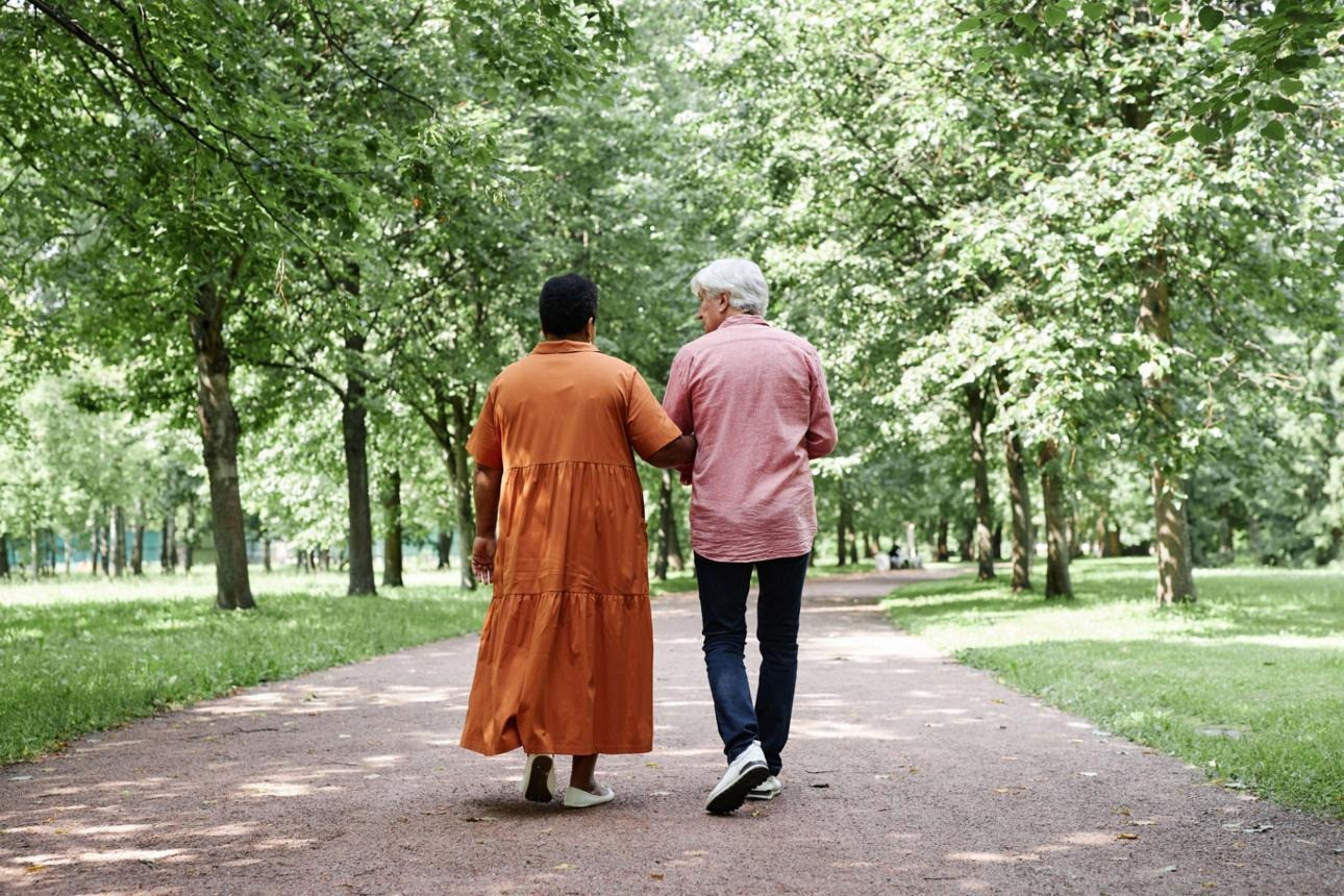 Older white-haired man in a pink shirt walking alongside a woman in an orange shirt, viewed from behind in a green outdoor setting.
