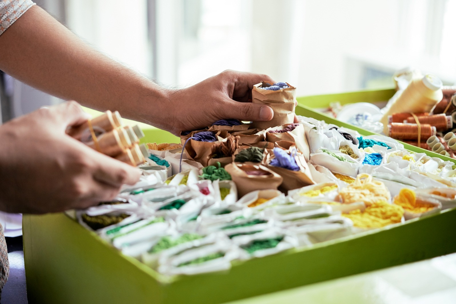Close-up of hands sorting colorful embroidery threads, yarn bundles, and sewing materials arranged in a green tray.