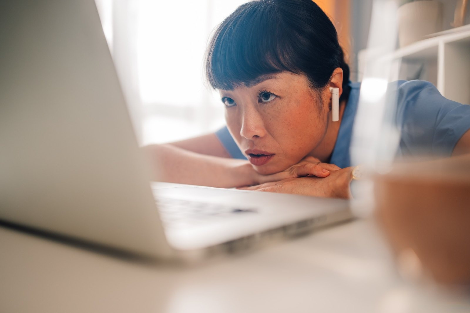 Woman wearing a blue shirt and wireless earbud looking attentively at a laptop while resting her chin on her hands.