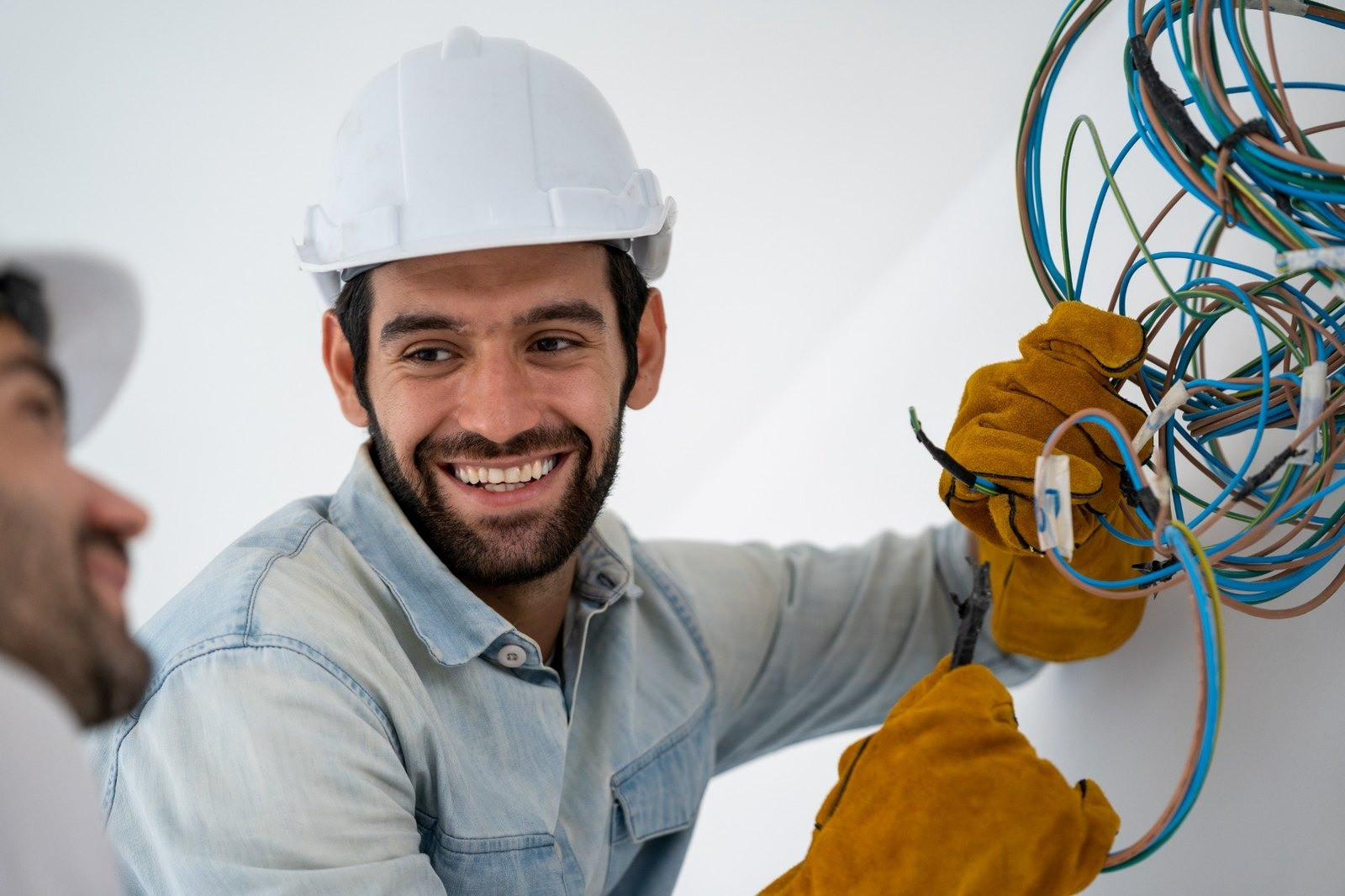 Electrician wearing a white safety helmet and yellow work gloves handling electrical wiring alongside a coworker.