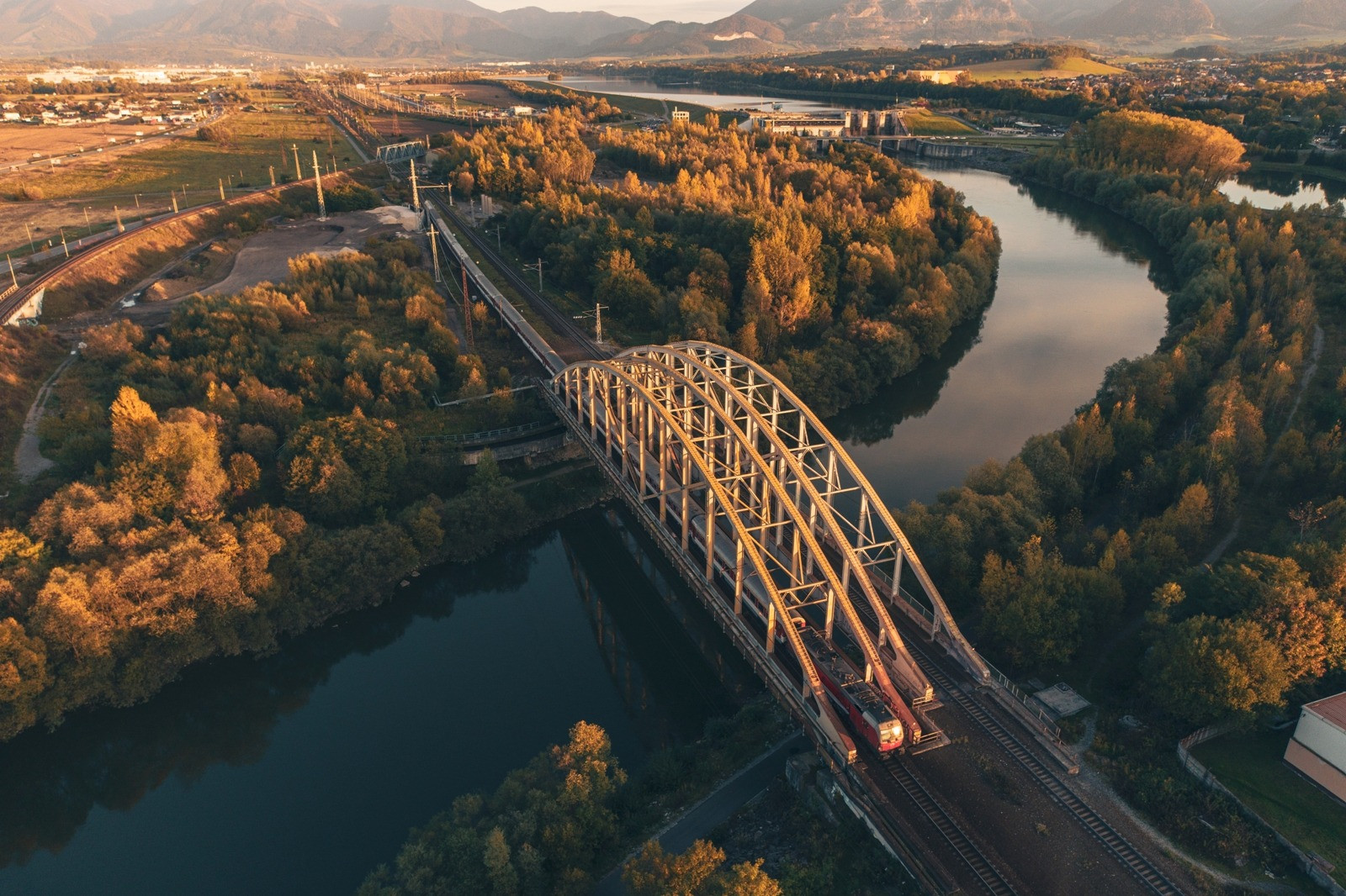 Aerial view of a train crossing a steel arch bridge over a river, surrounded by golden trees and distant mountains.