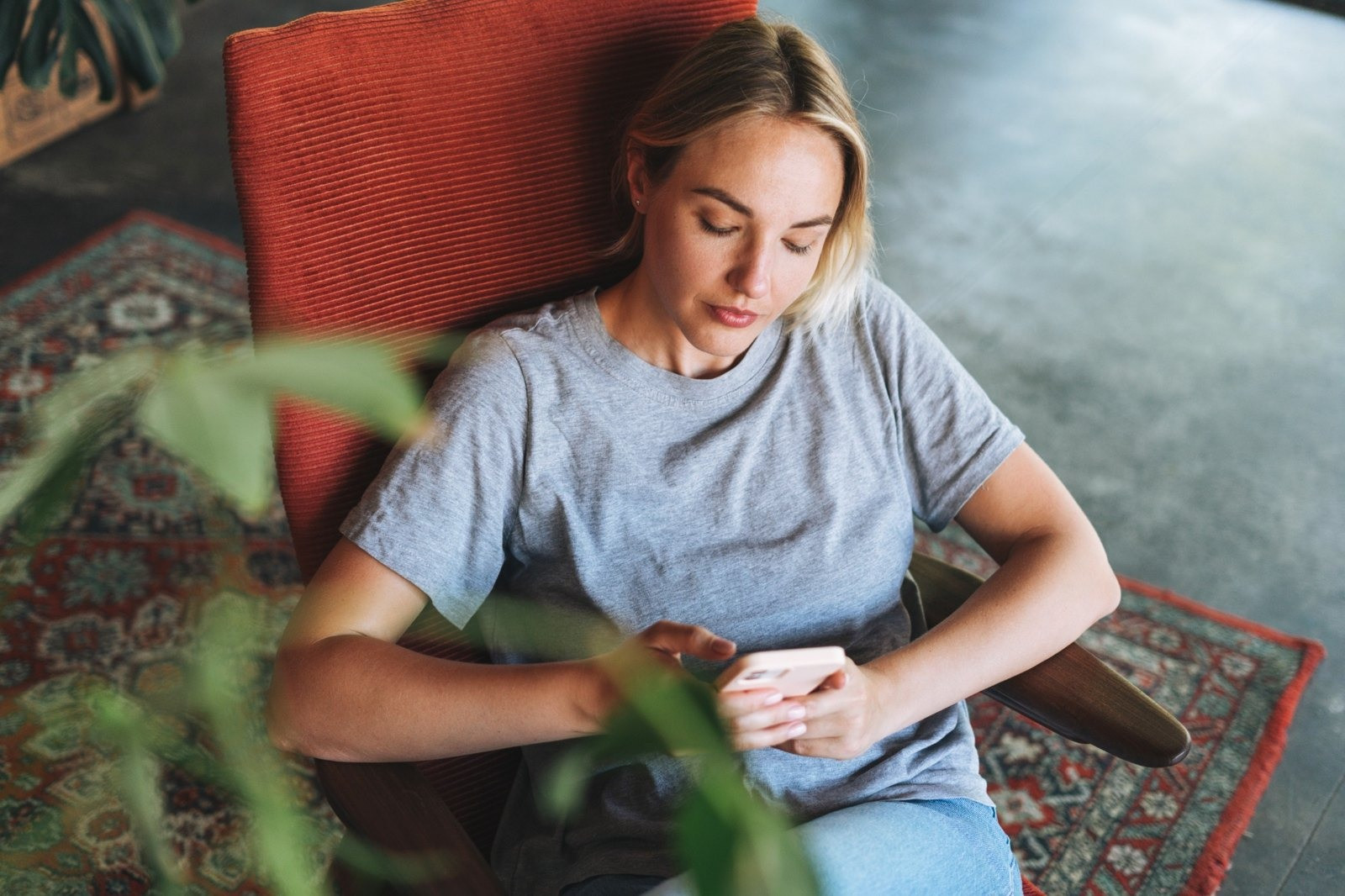 Blonde woman sitting in a red chair and looking at her smartphone in a quiet indoor setting.