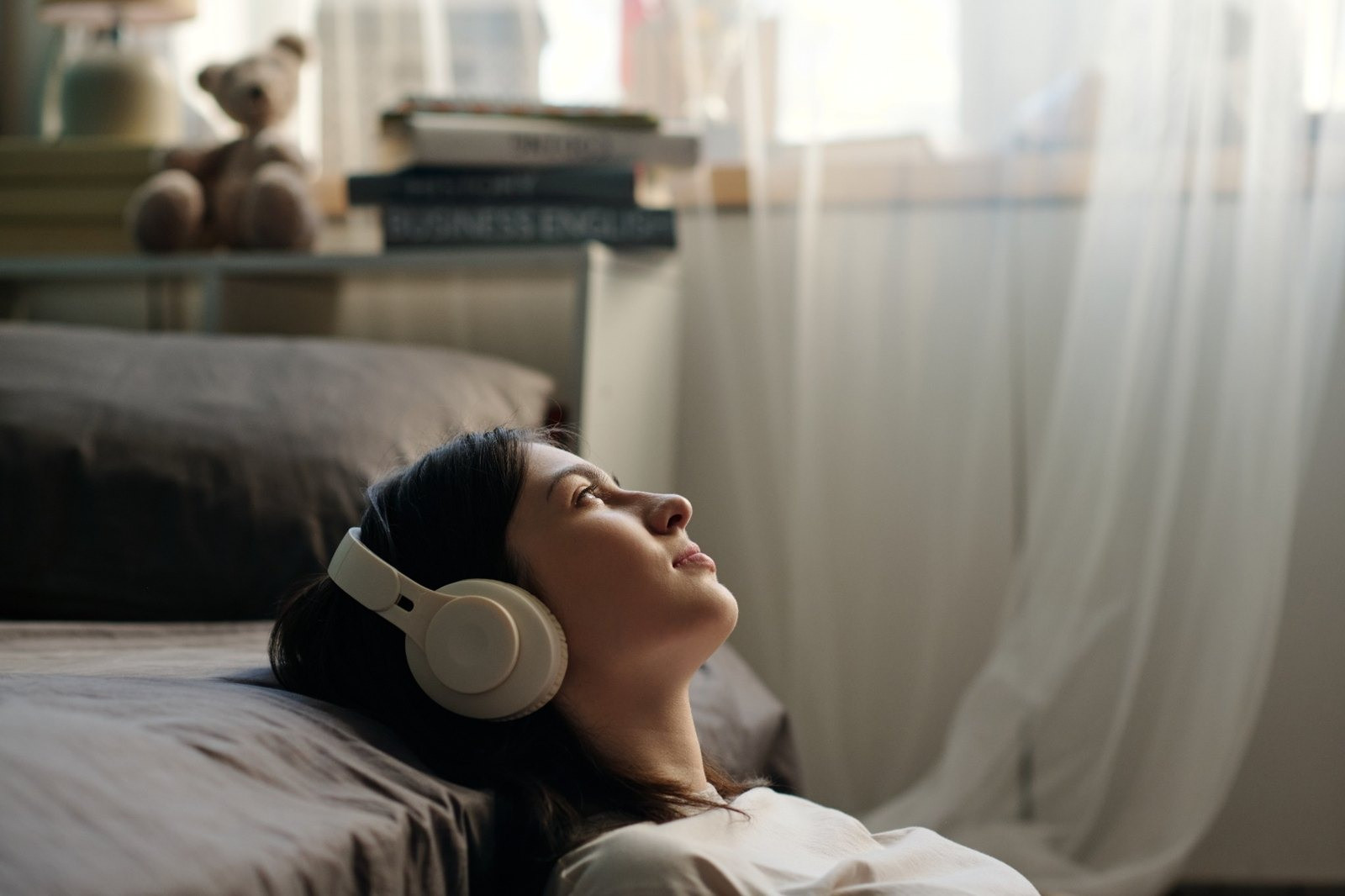 Woman lying on a bed wearing headphones in a softly lit bedroom.