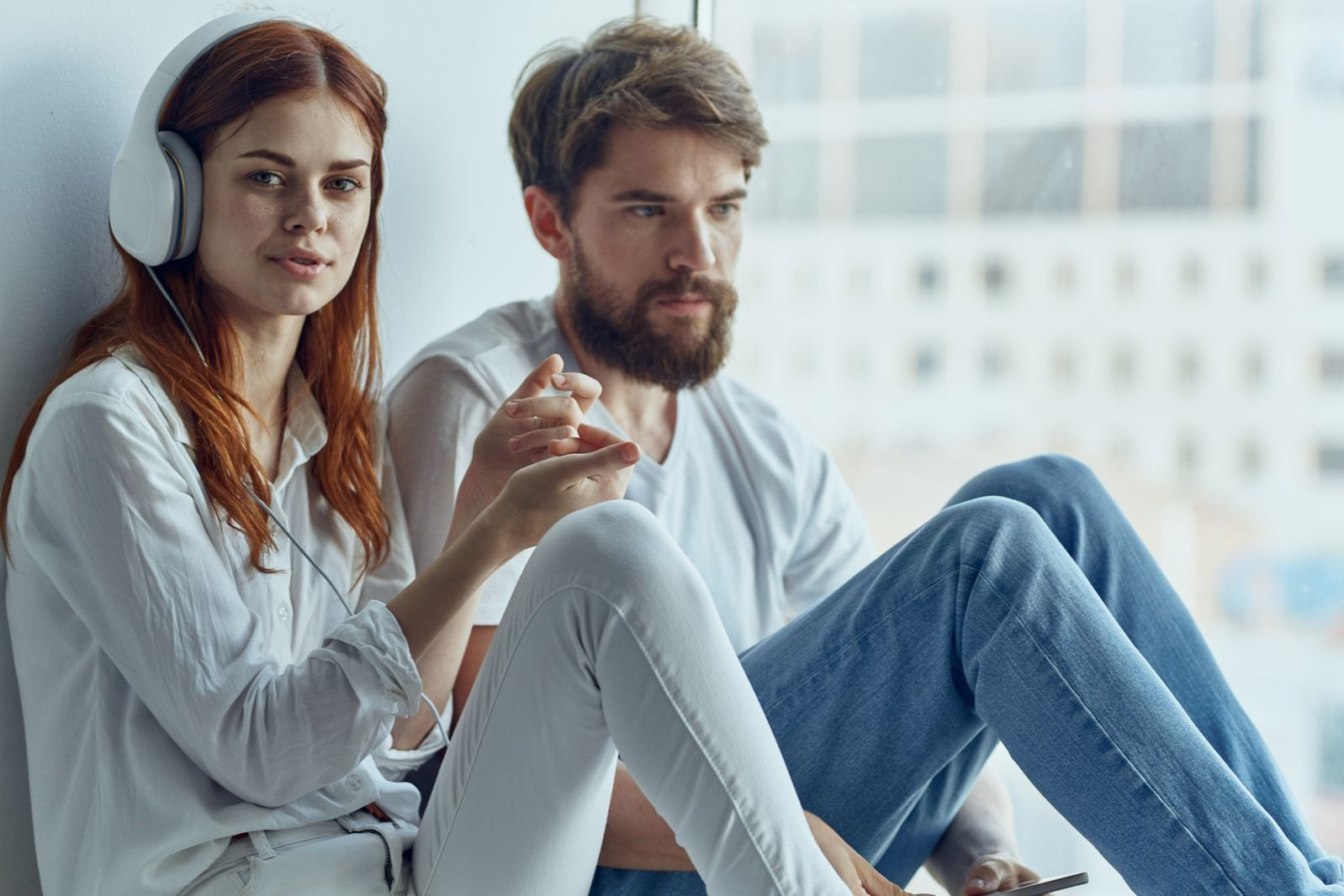 Woman wearing headphones sitting beside a man near a window, both appearing thoughtful and relaxed.