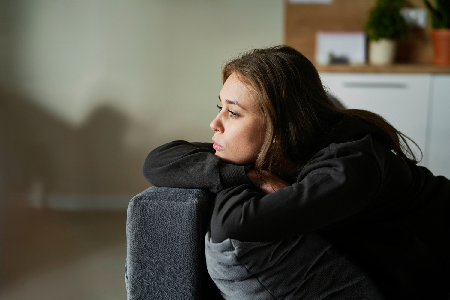 Young woman sitting curled up on a sofa, looking thoughtful in a quiet home setting.