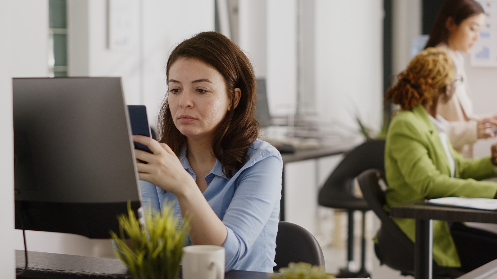 Woman sitting at a desk in an office, looking at her phone beside a computer while appearing slightly distracted.