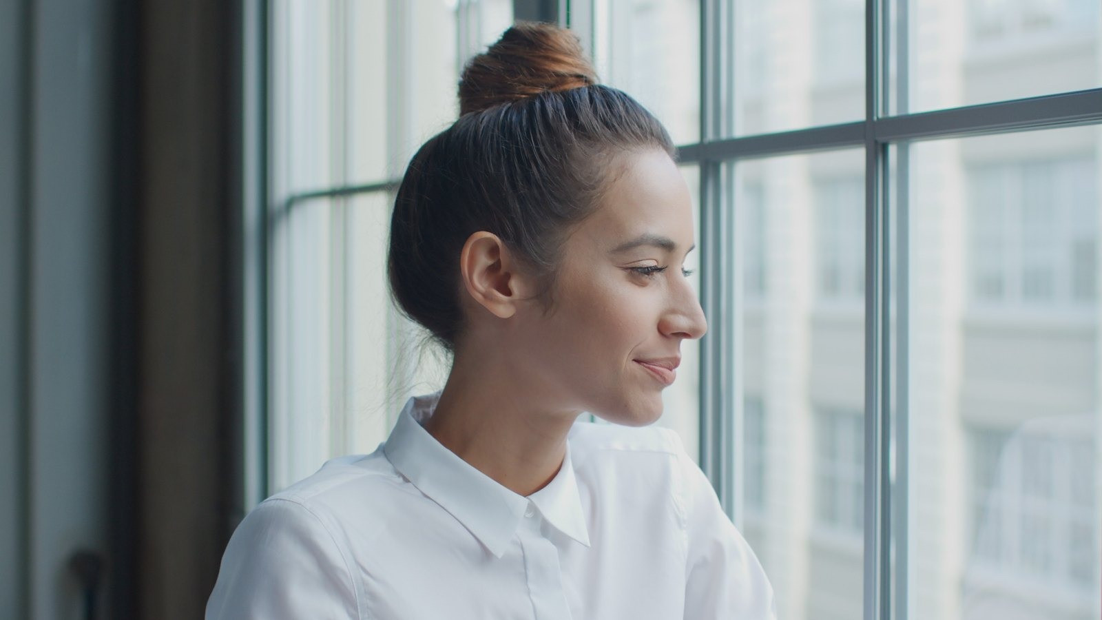 Woman with her hair in a bun wearing a white blouse and looking thoughtfully out a window.