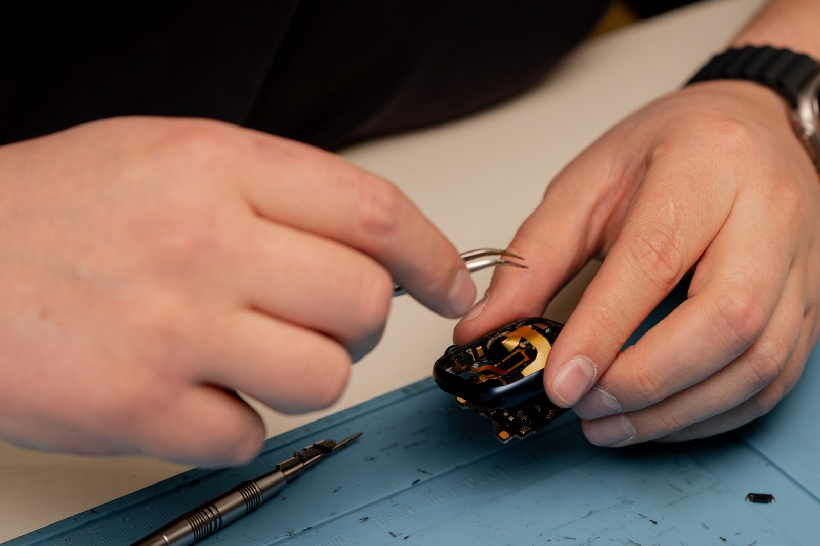 Close-up of a technician using precision tweezers to repair a small electronic device on a blue work mat, with a screwdriver nearby.