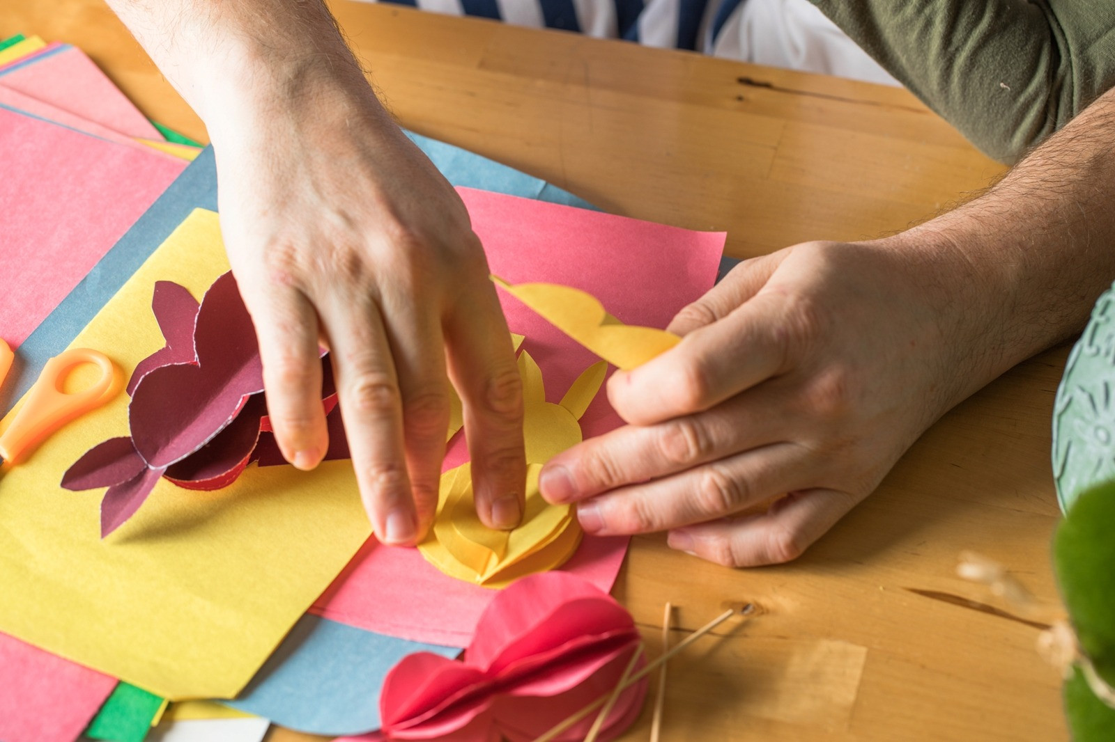 Close-up of hands arranging colorful paper cutouts and craft materials on a wooden table during a creative activity.
