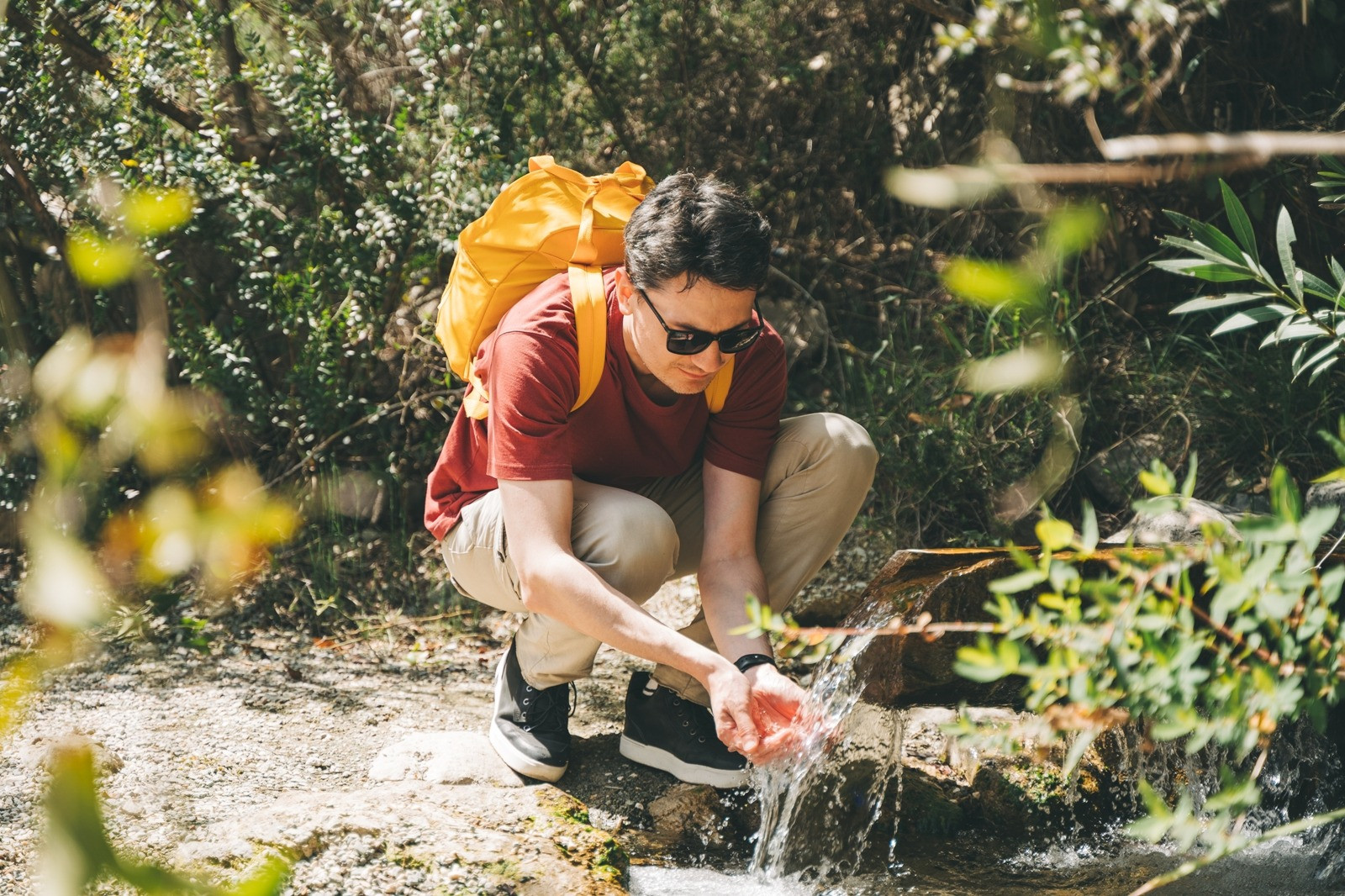 Man with a backpack crouching beside a natural spring and washing his hands in flowing water during a hike.