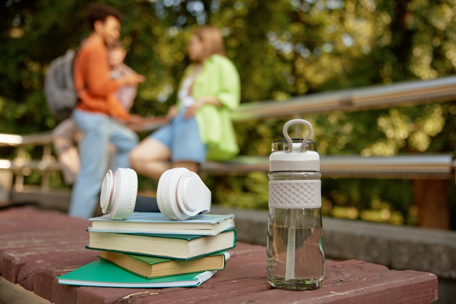 White headphones resting on stacked books beside a water bottle on an outdoor table, with people blurred in the background.