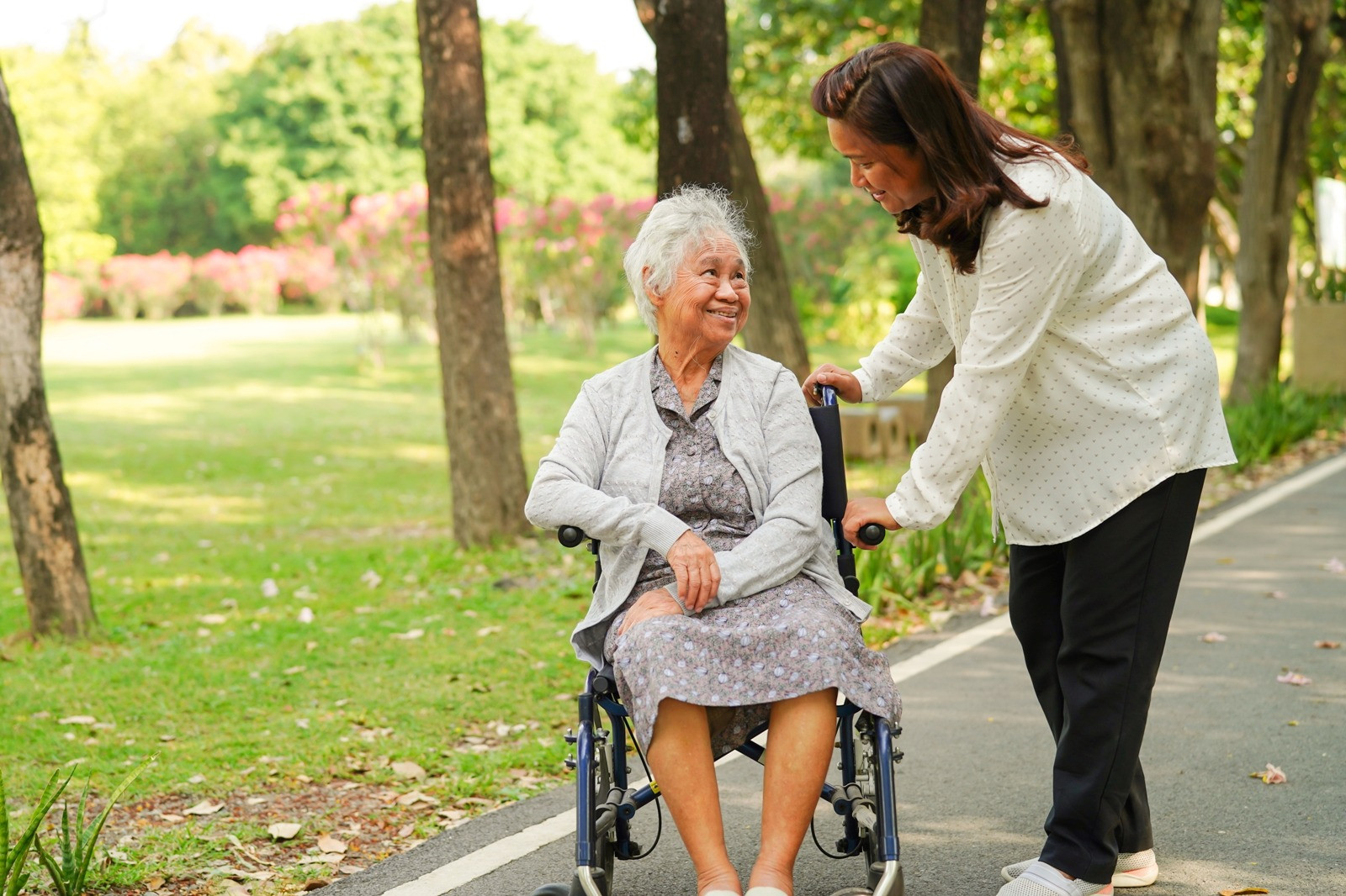 Older woman in a wheelchair smiling while another woman assists her outdoors in a peaceful park.