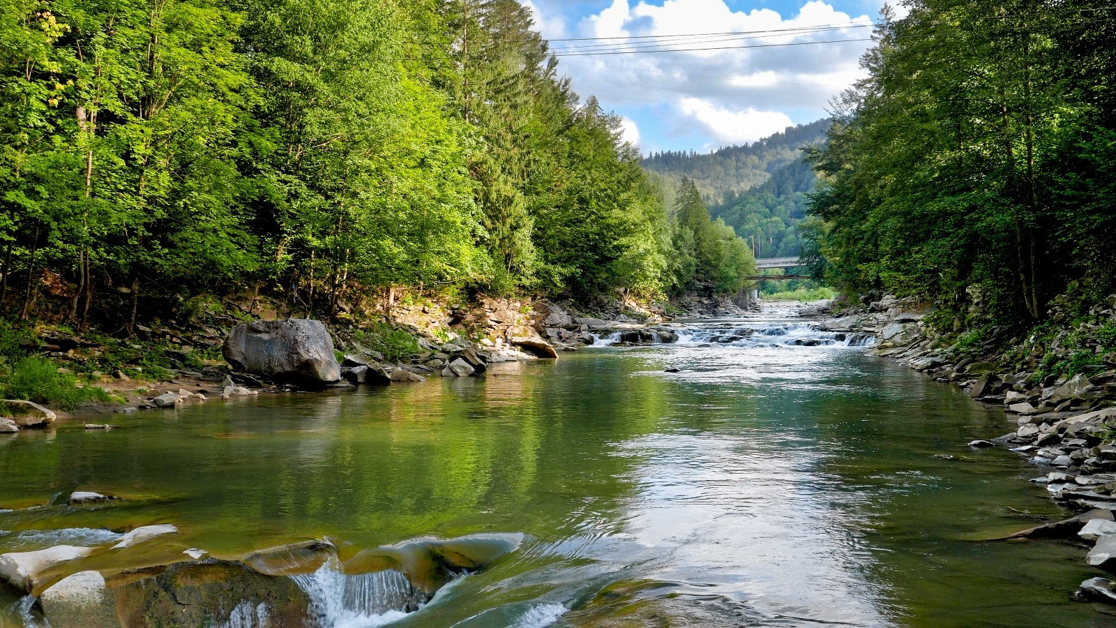 A river flows over rocks between lush green forest banks, with wooded hills and a partly cloudy sky in the distance.