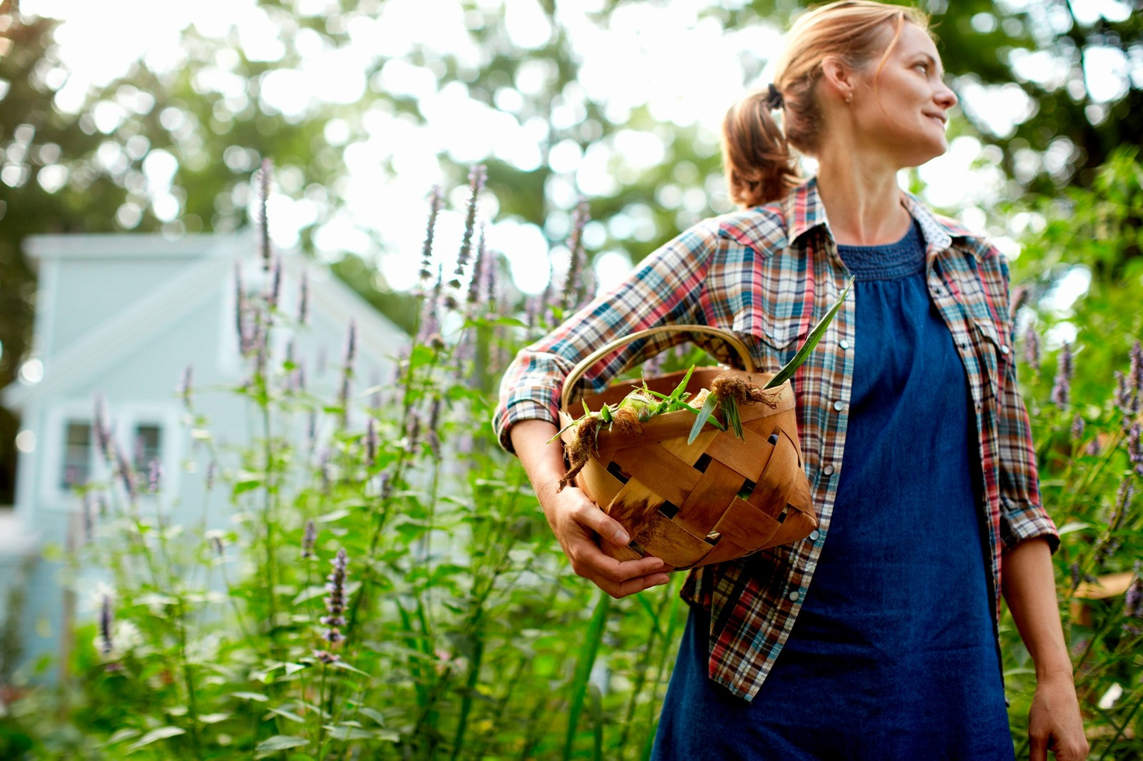 Woman in a plaid shirt holding a woven basket of freshly harvested vegetables while standing in a lush garden surrounded by greenery and purple flowers.