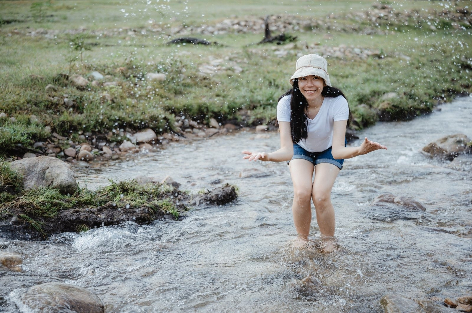 Smiling woman wearing a beige bucket hat, white shirt, and denim shorts while splashing water in a shallow stream.