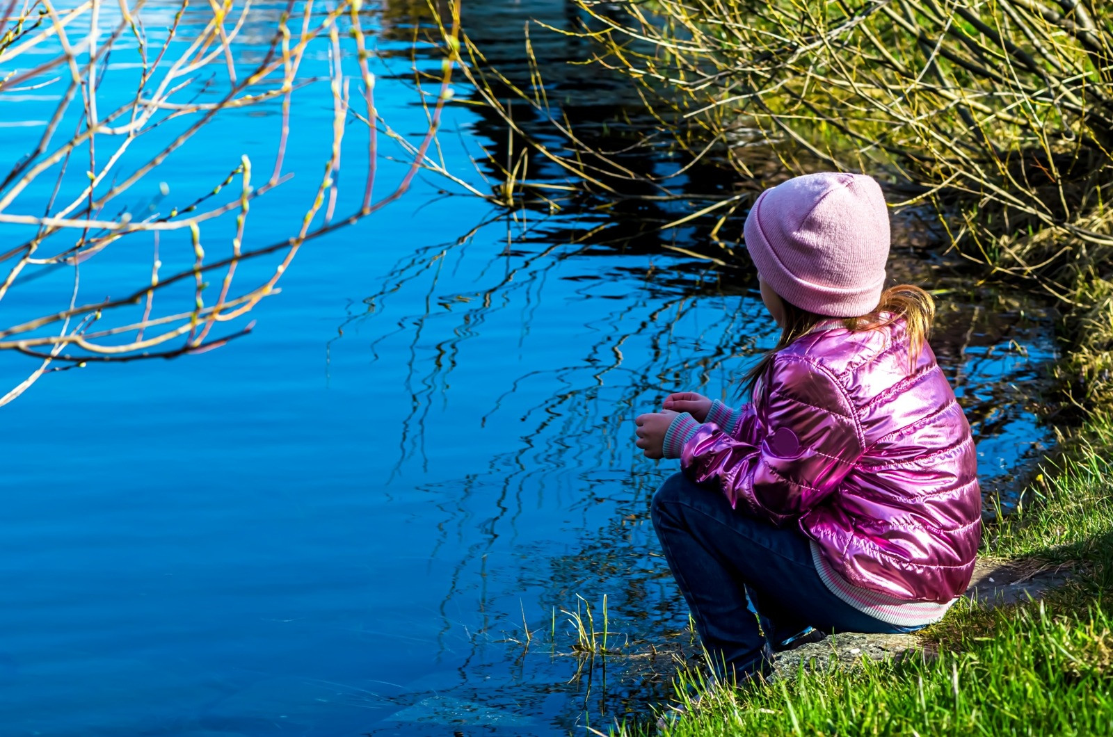 Person sitting beside a calm pond, looking at the water in a quiet outdoor setting.