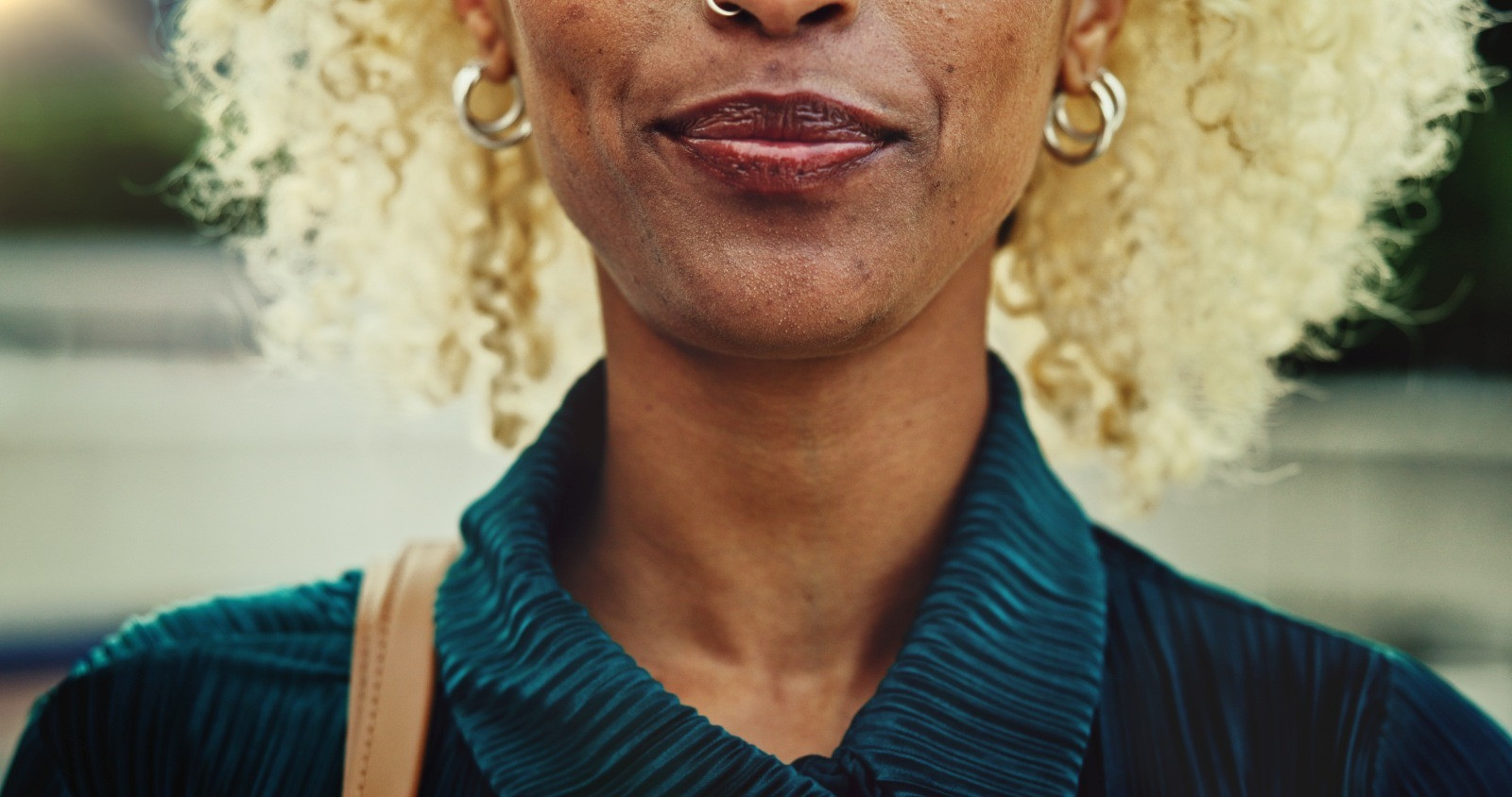 Close-up of a woman’s lower face and shoulders, with blonde curls, silver hoop earrings, and a teal collared top.
