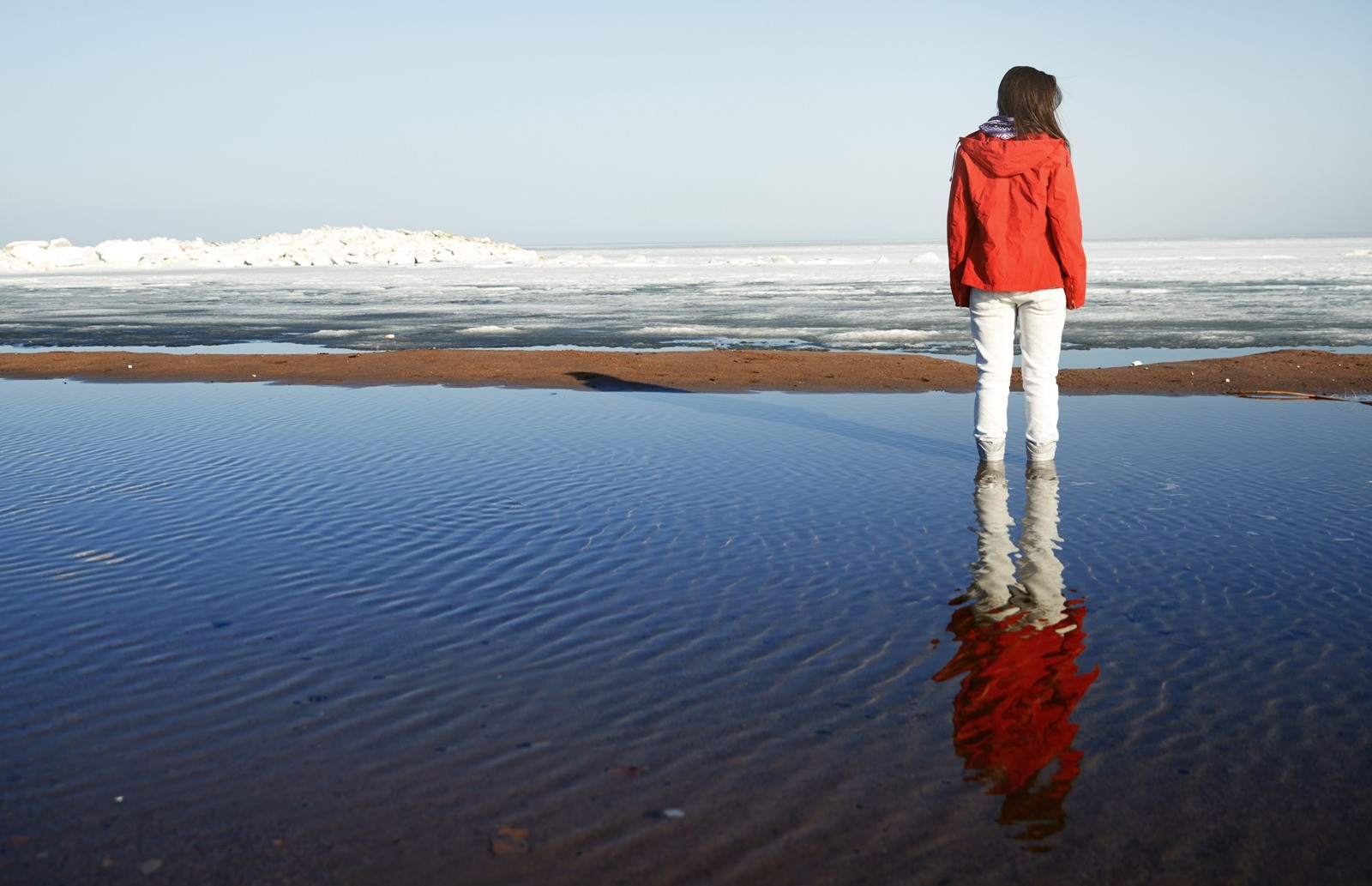 Woman in a red jacket standing in shallow water, looking toward a calm, icy shoreline.